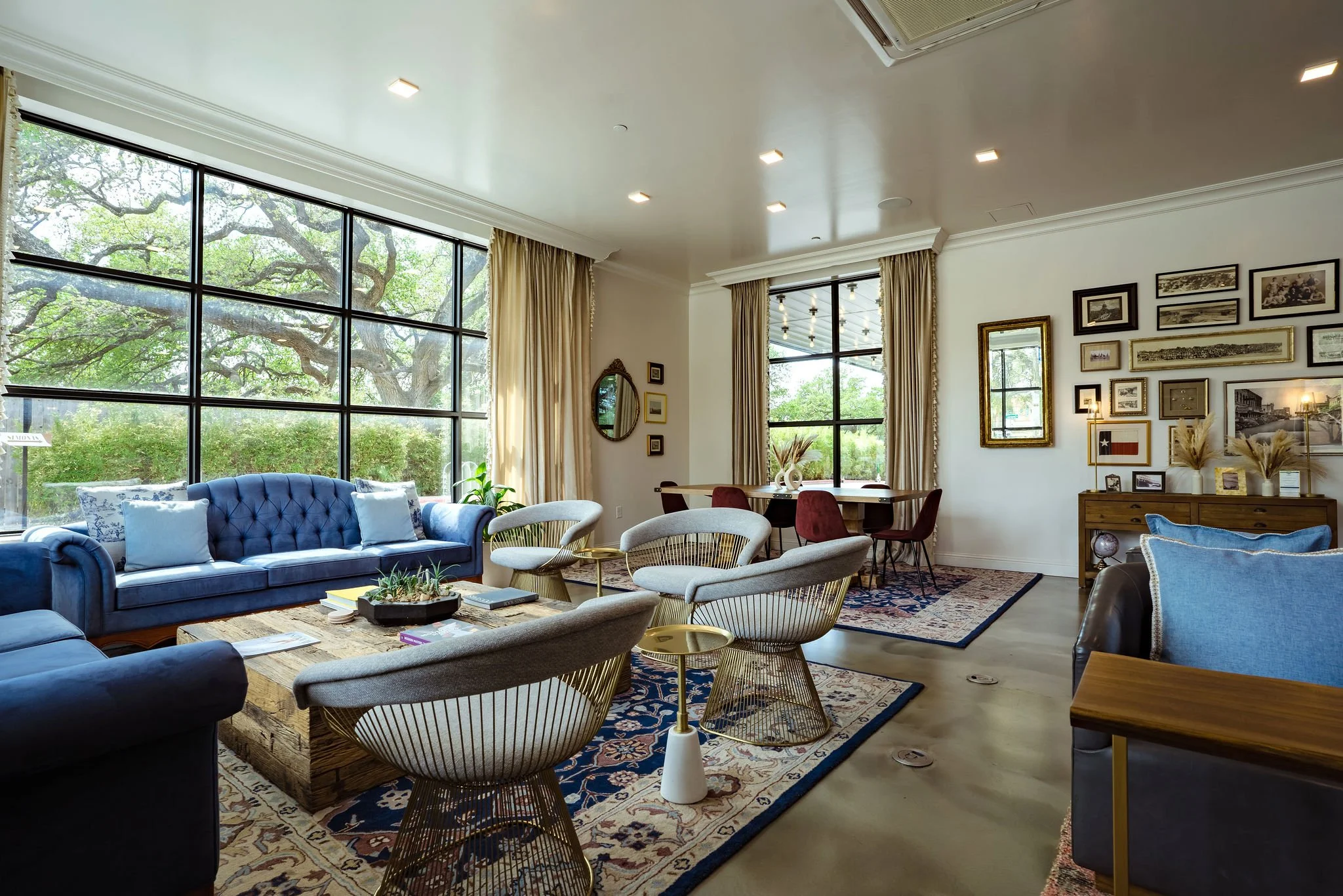 Spacious living room with large windows, blue velvet sofa, beige chairs, wooden coffee table, and a dining area with chairs near large windows, decorated with framed pictures and mirrors on the walls, and various plants and decorative items.