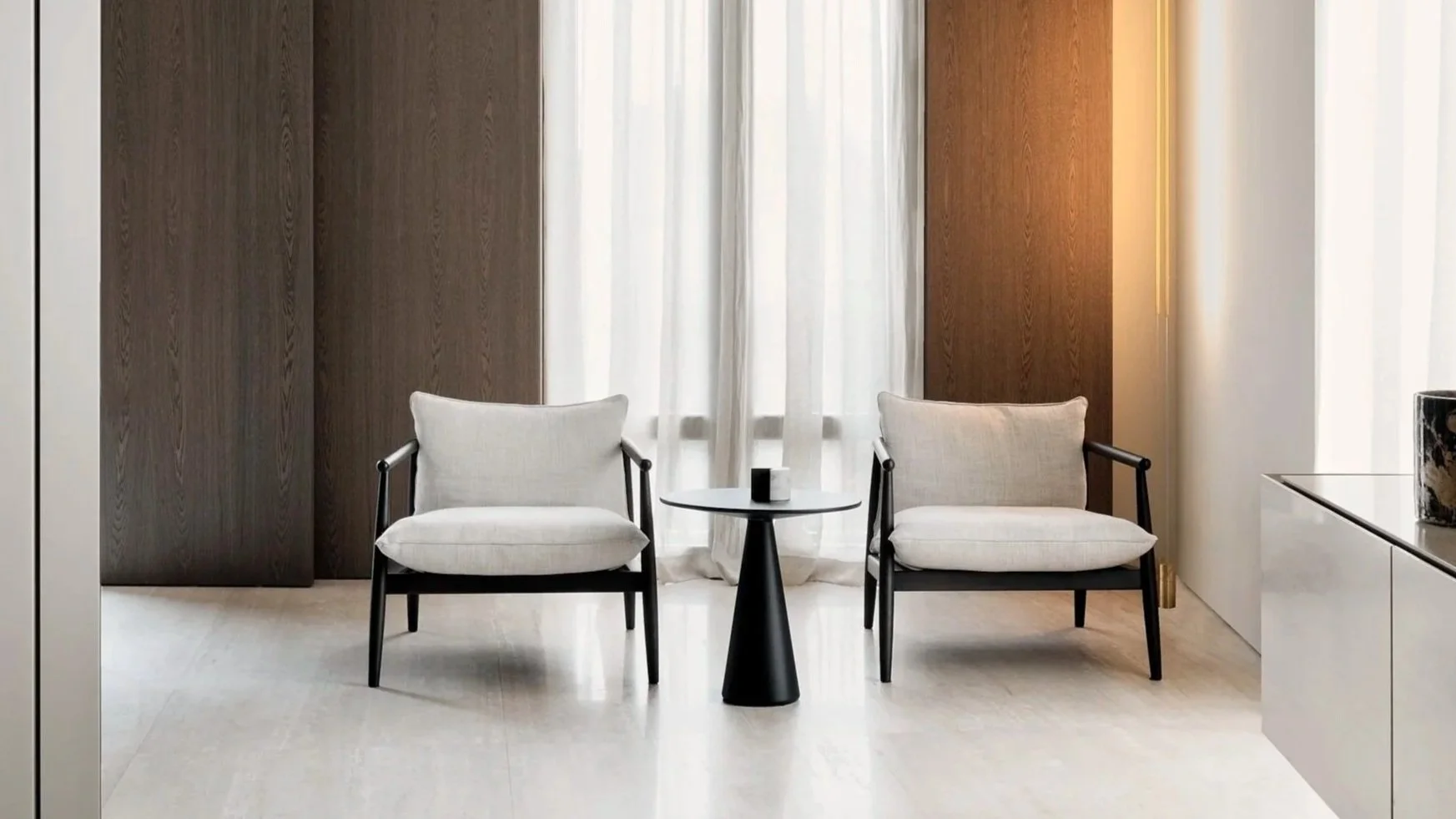 A modern interior with two white armchairs with black legs and armrests, a small round black table between them, behind which are white curtains on the left side, and a wooden panel on the right side, with partial view of a white cabinet and a floral