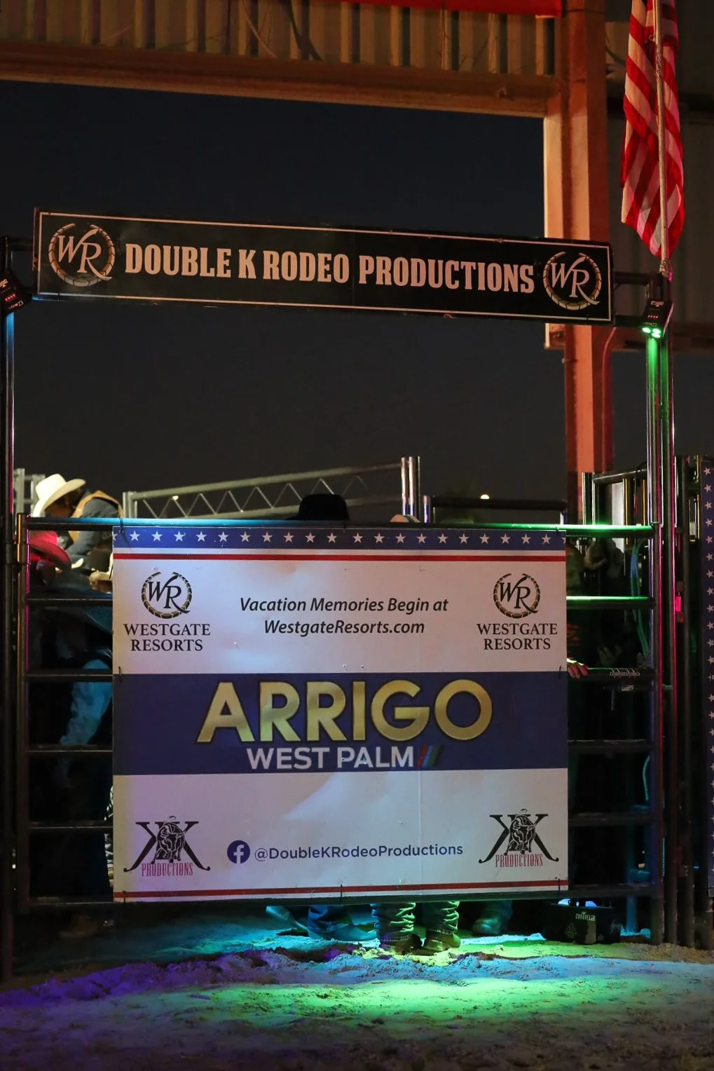 Photo of a rodeo arena entrance with signs for Double K Rodeo Productions and Arrigo West Palm, promoted by Westgate Resorts, at night. There is an American flag hanging to the right.