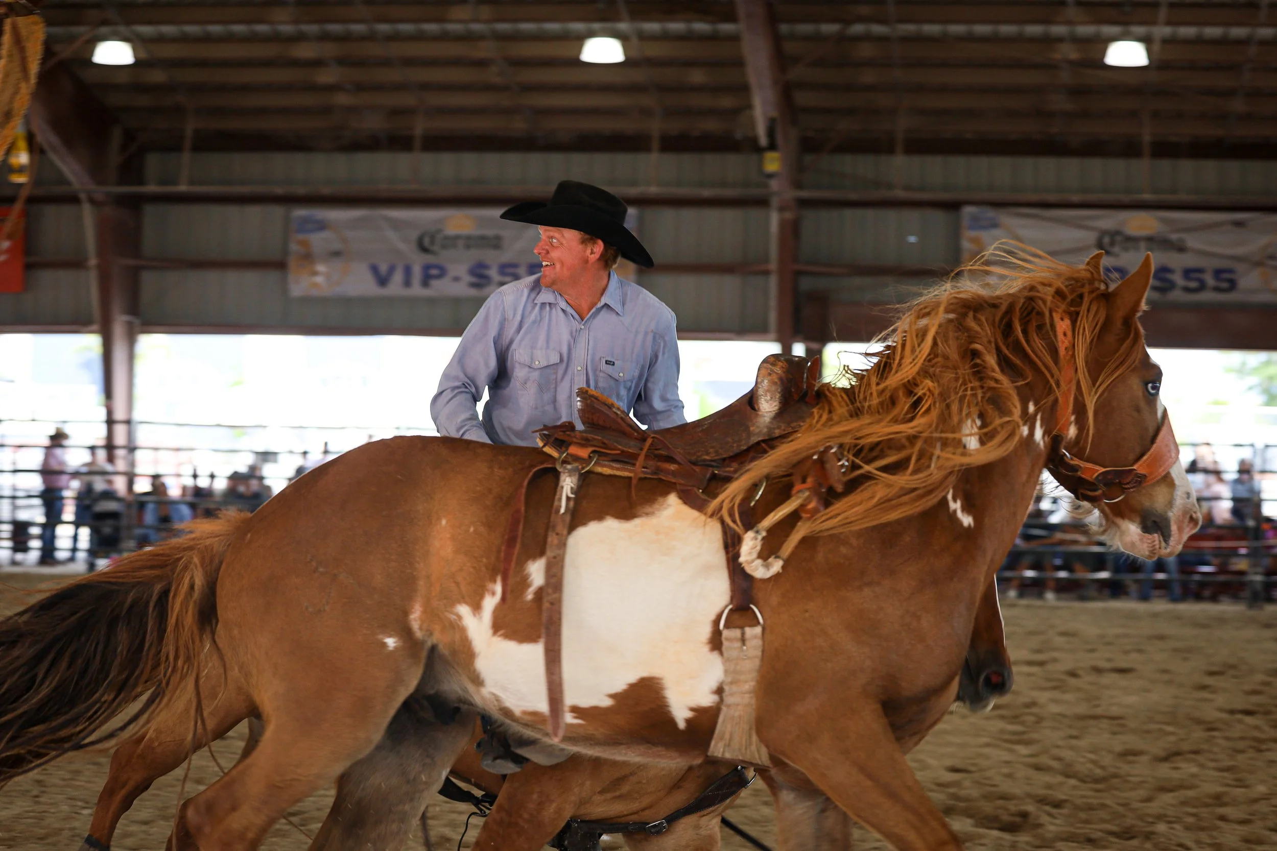 A man wearing a cowboy hat riding a galloping chestnut and white paint horse inside an indoor rodeo arena.
