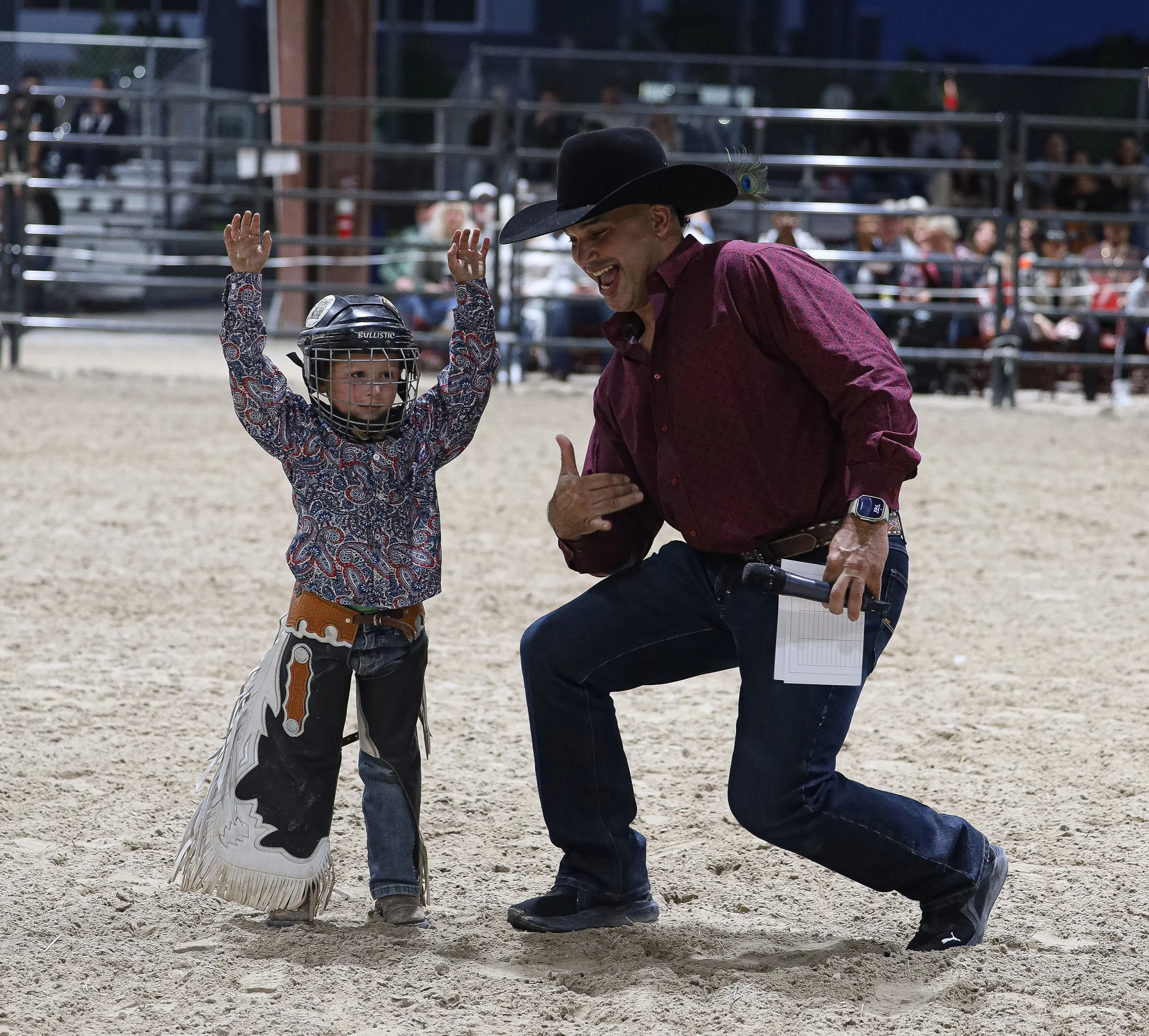A young child wearing a cowboy hat and protective helmet celebrates with an adult man dressed in Western attire at a rodeo arena. The child has raised arms, and the man is kneeling and smiling while holding a notepad and pointing to the child.