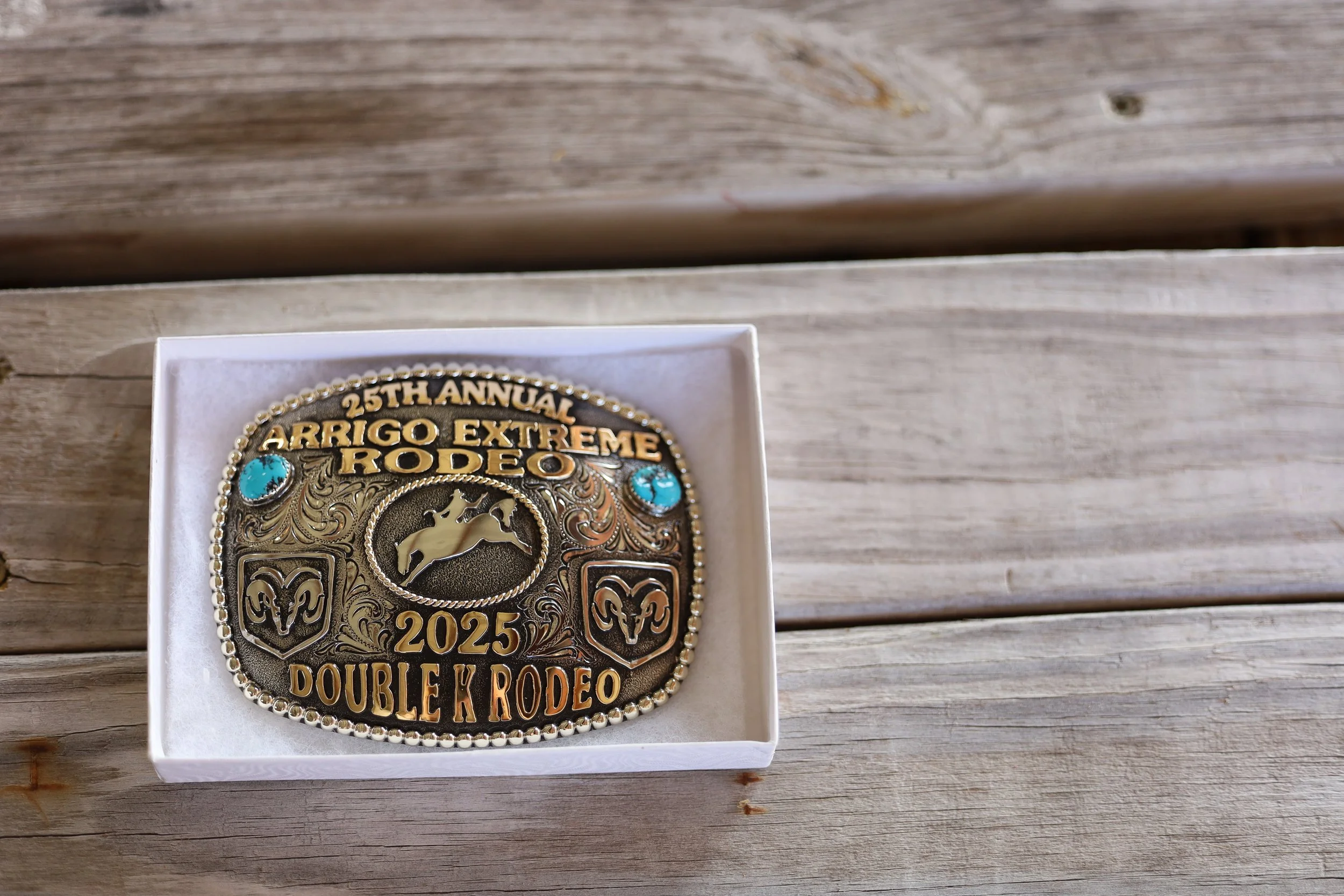 A commemorative belt buckle for the 25th Annual Arrigo Extreme Rodeo 2025 Double K Rodeo, placed in a white box on a wooden surface.