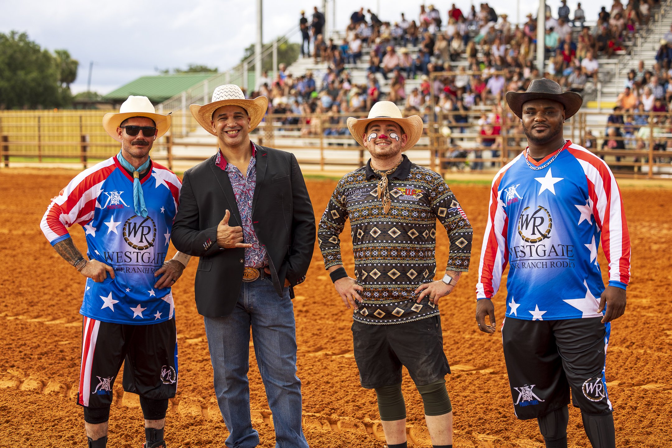 Four men standing on a dirt rodeo arena, with a crowd of spectators seated on bleachers in the background.