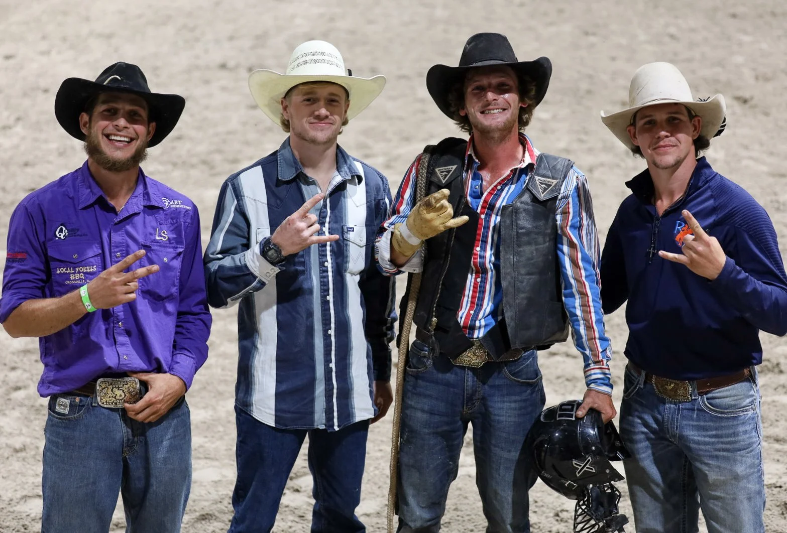 Four men wearing cowboy hats and jeans standing on dirt ground, smiling and posing for the photo, with some making rock and roll hand signs.
