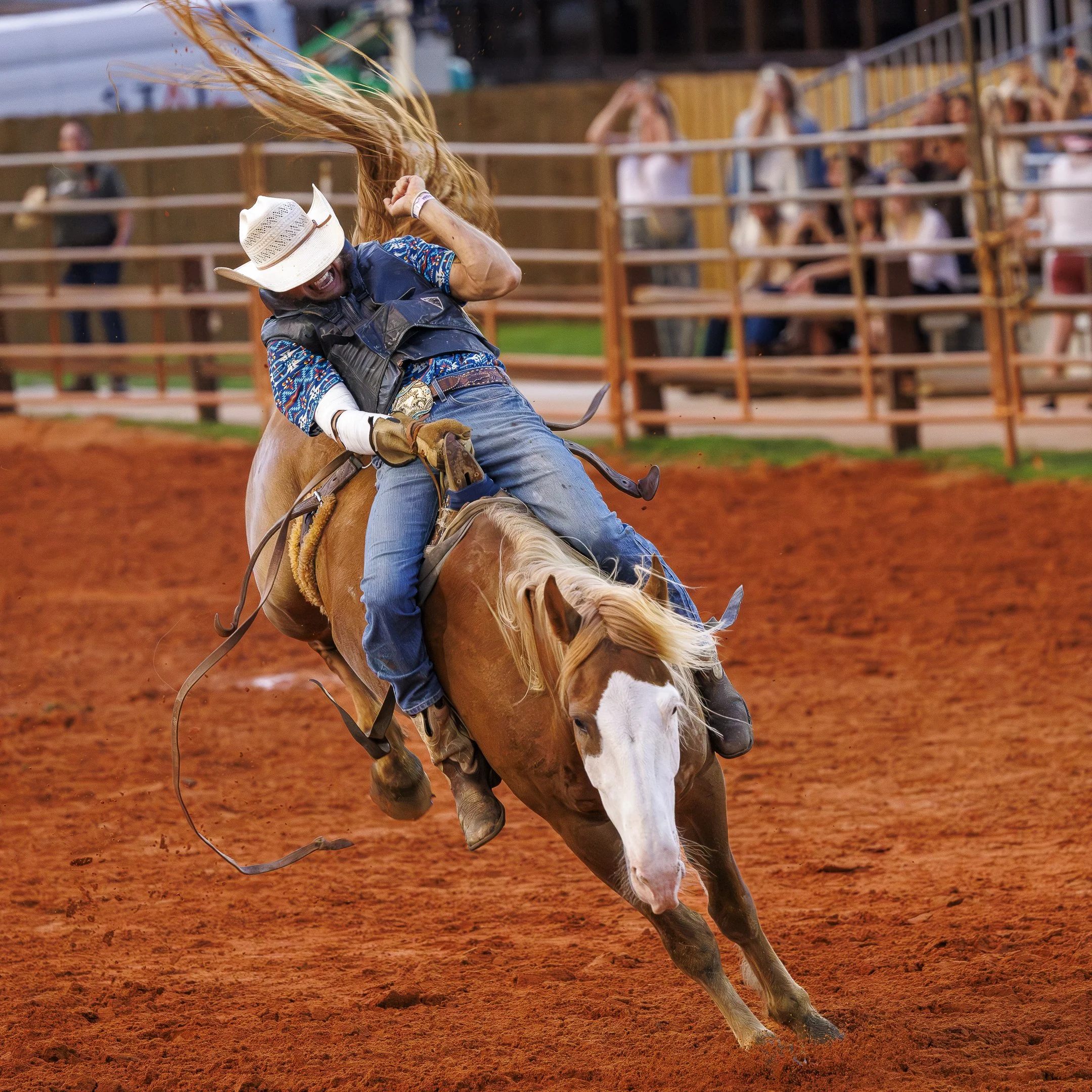 A cowboy in a cowboy hat and western attire riding a bucking horse on a dirt rodeo arena, with spectators watching from the stands.