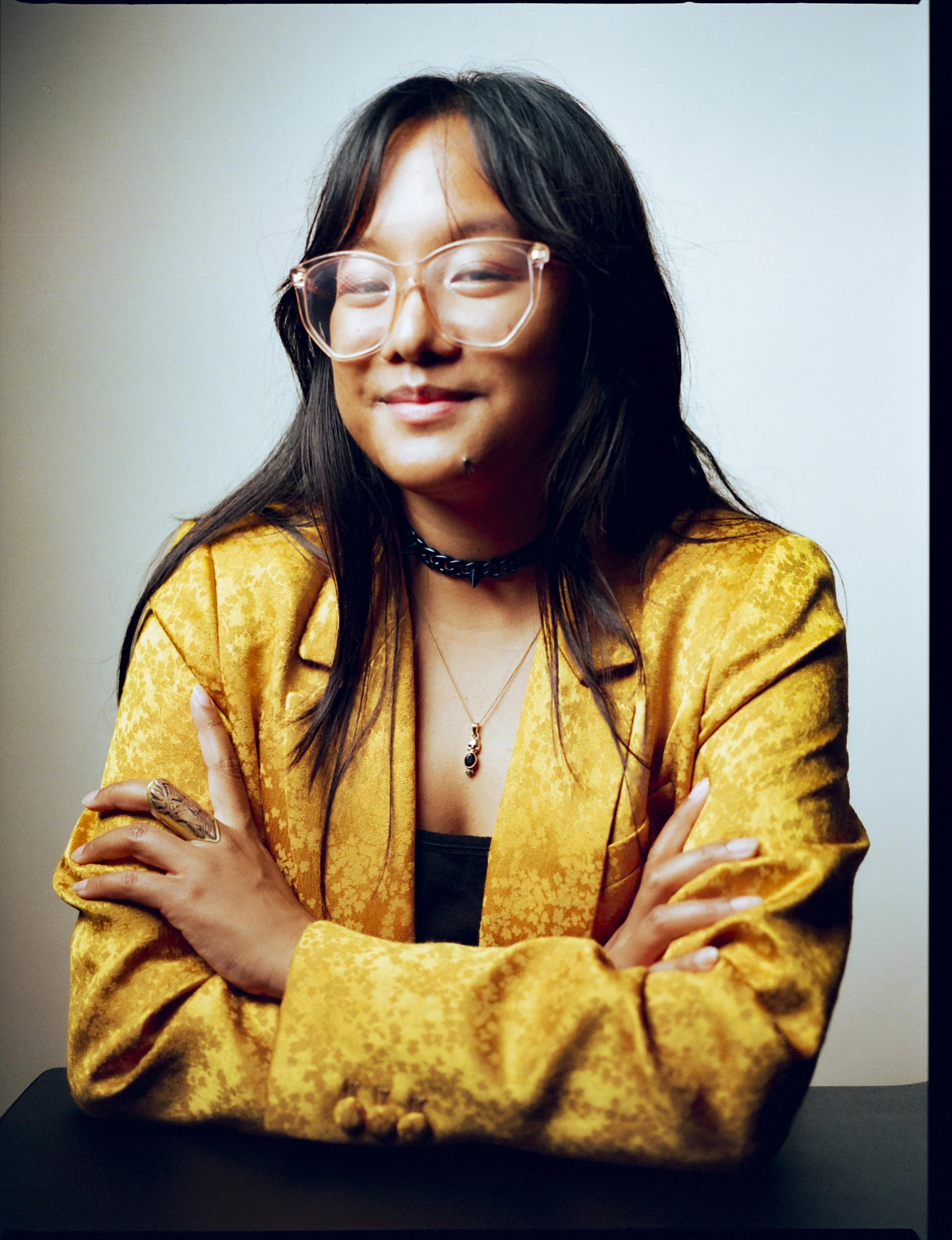 A brown femme with geometric glasses, in a bright golden yellow blazer and dark blue choker, grins gently, arms folded on a low black table in front of them.
