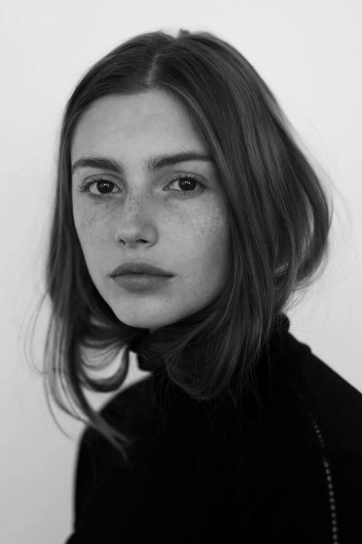 Black and white close-up portrait of a young woman with shoulder-length hair, freckles, and a super short turtleneck top, looking directly at the camera.