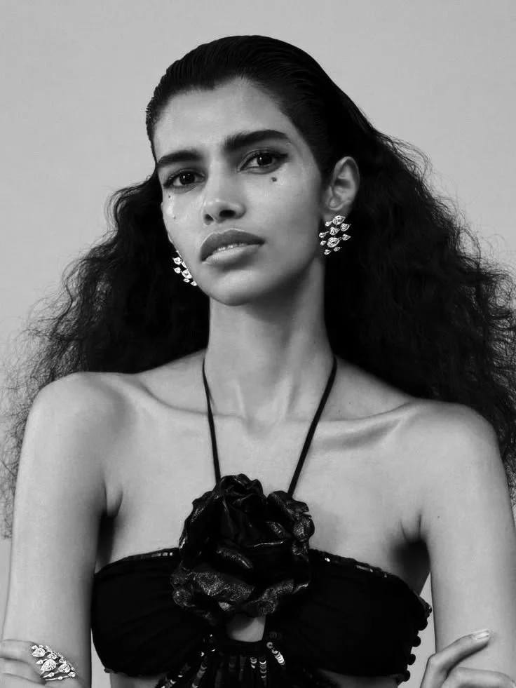 Black and white portrait of a woman with curly hair, wearing earrings, a necklace with a large flower pendant, and a strapless top with a floral embellishment.