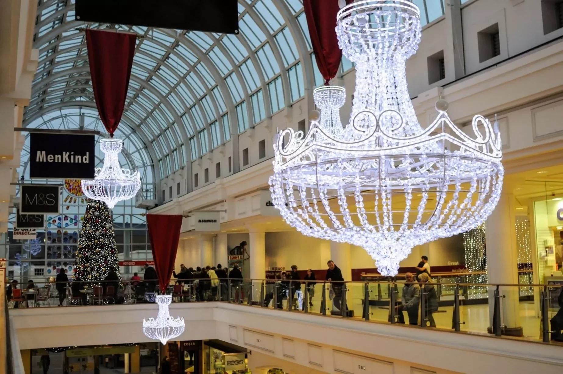 Large chandelier-style Christmas lights hanging inside the Royal Priors Shopping Centre in Leamington Spa, with festive decorations and shoppers in the background.