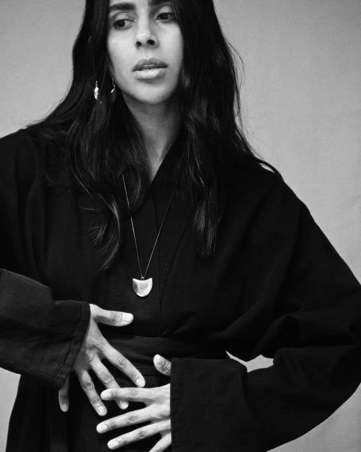 A black-and-white photo of a woman with long dark hair, wearing a dark top and a necklace with a crescent-shaped pendant, looking to her left with a serious expression, holding her stomach with her left hand.