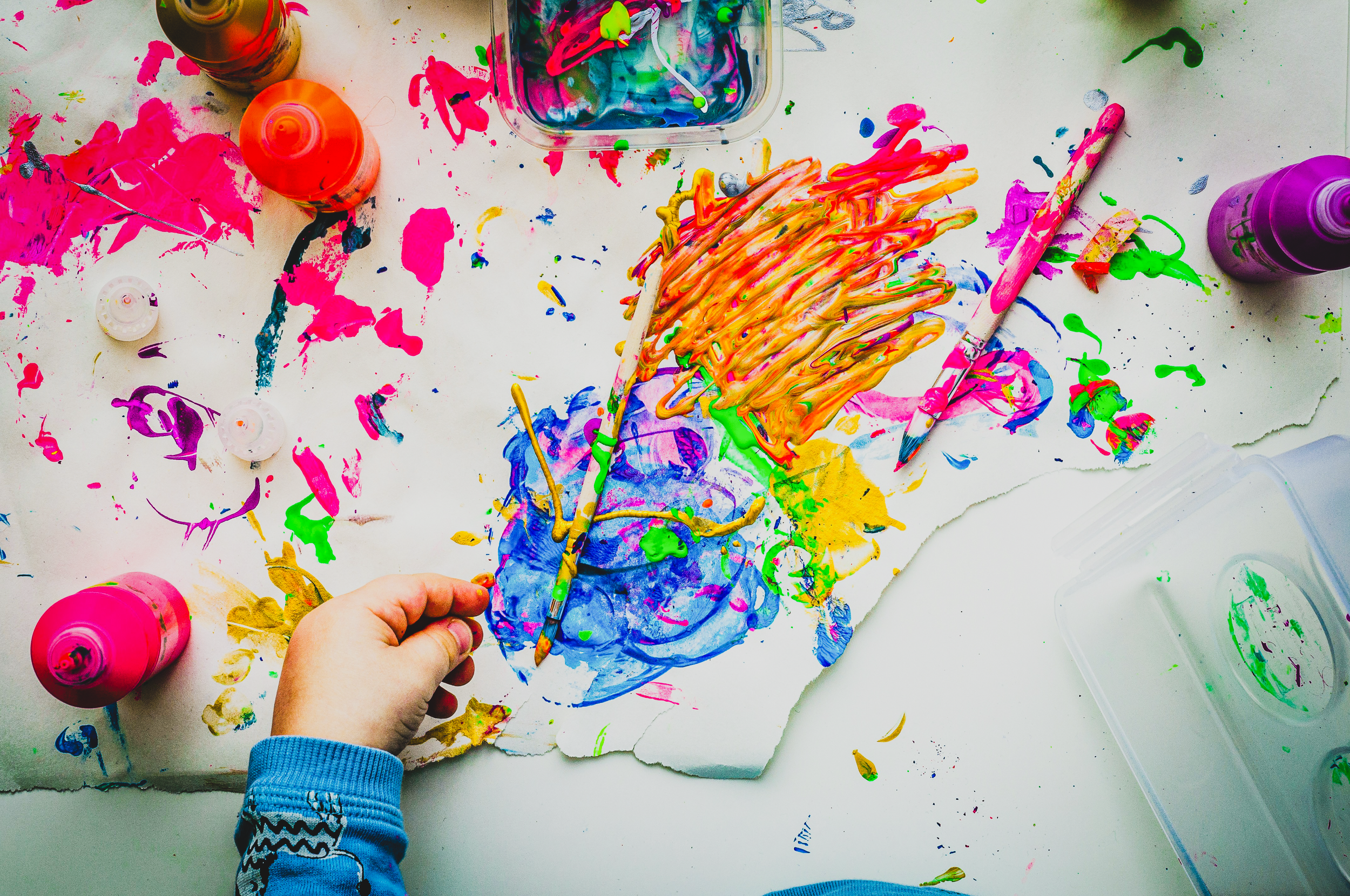 Photo by: Dragos Gontariu, a small child's hand with bright paint splatters and art supplies on table