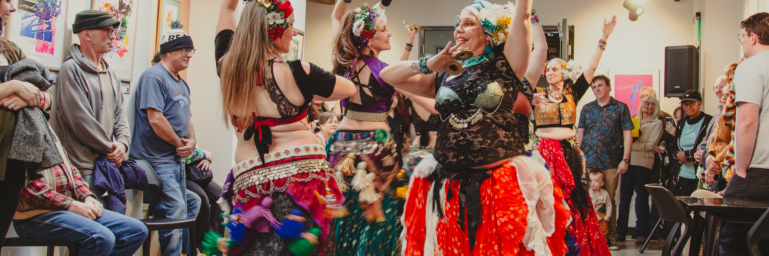 Belly dancers in colorful skirts perform for a crowd standing in an art gallery