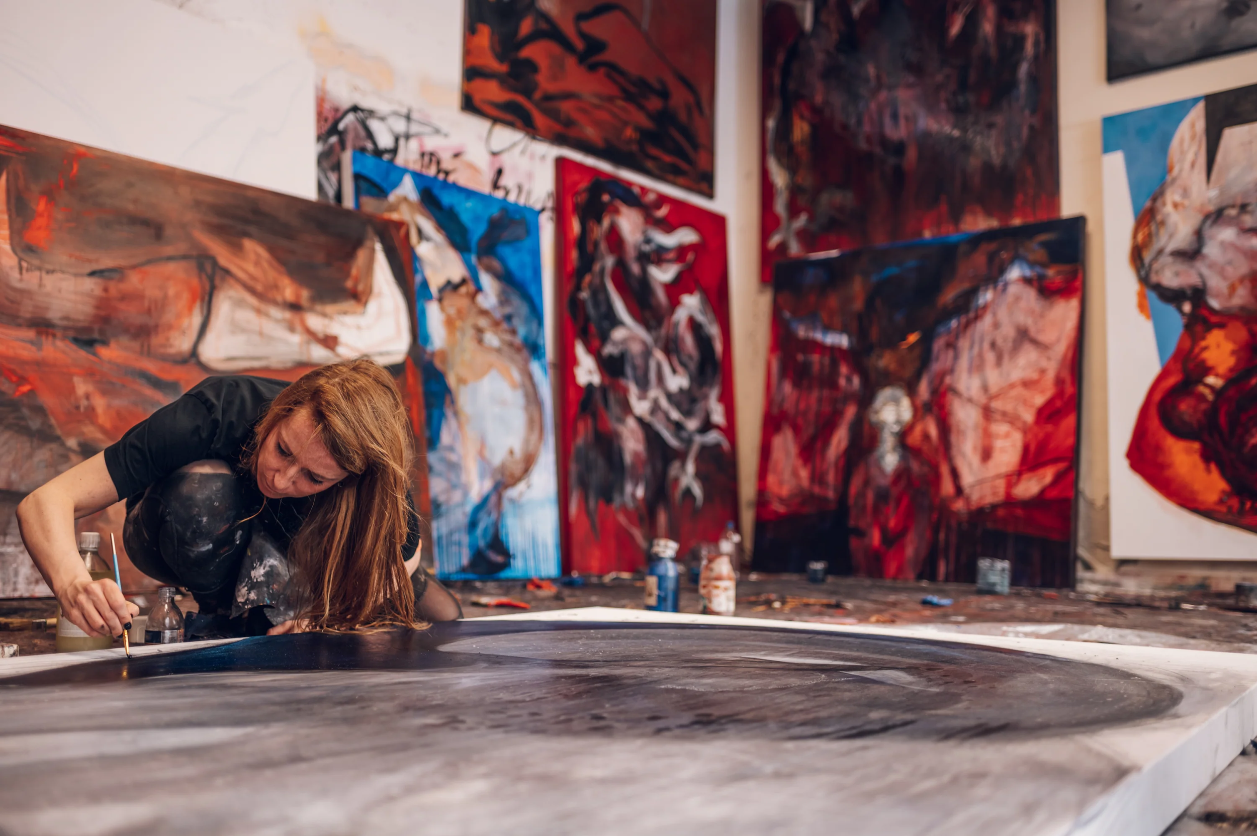 Artist kneeling down over a large painting with several larger abstract works on the walls behind her