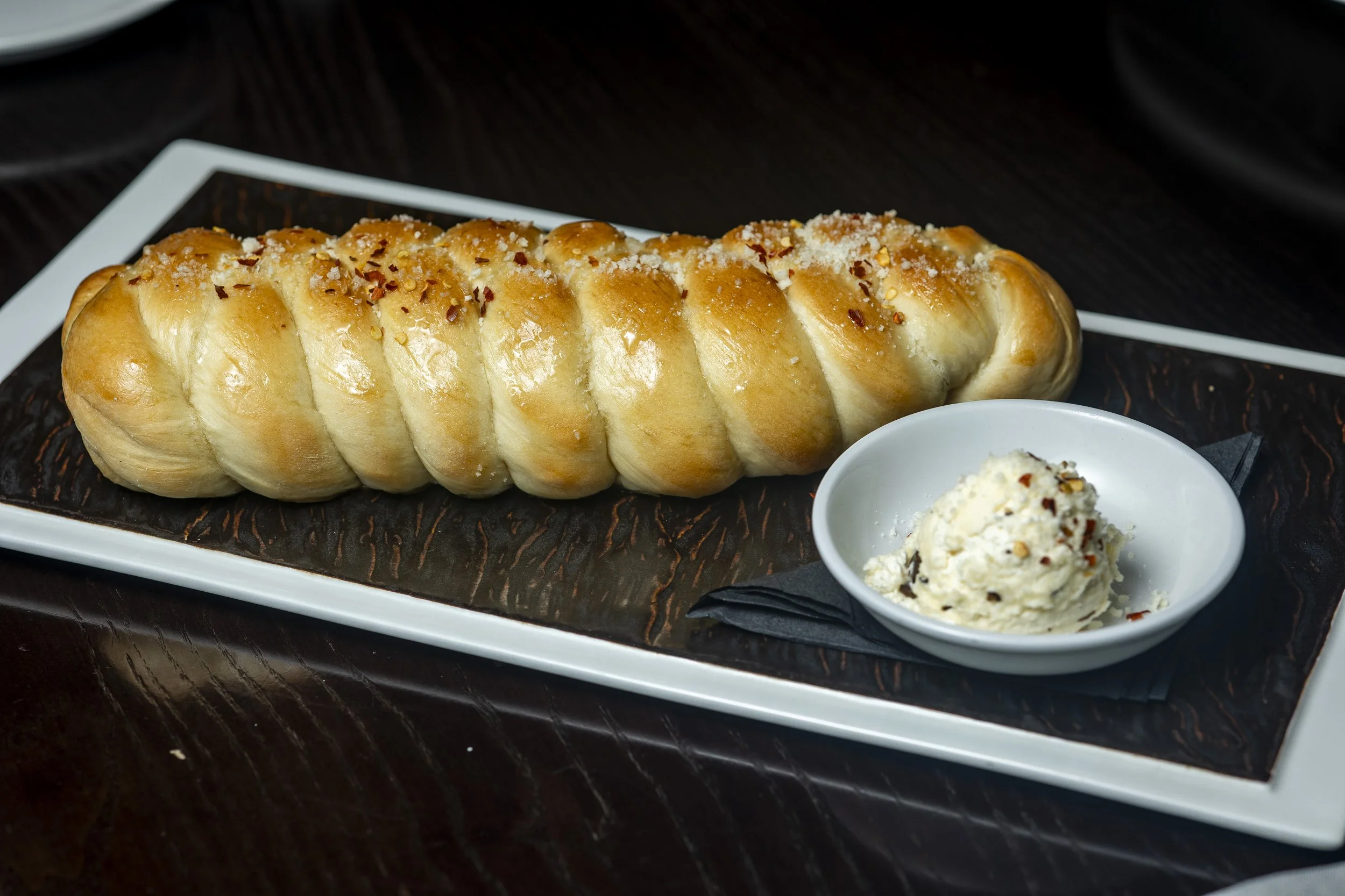 A long, golden-brown braided bread loaf topped with grated cheese and red pepper flakes on a white rectangular platter, with a small bowl of butter or soft cheese garnished with red pepper flakes on the side.