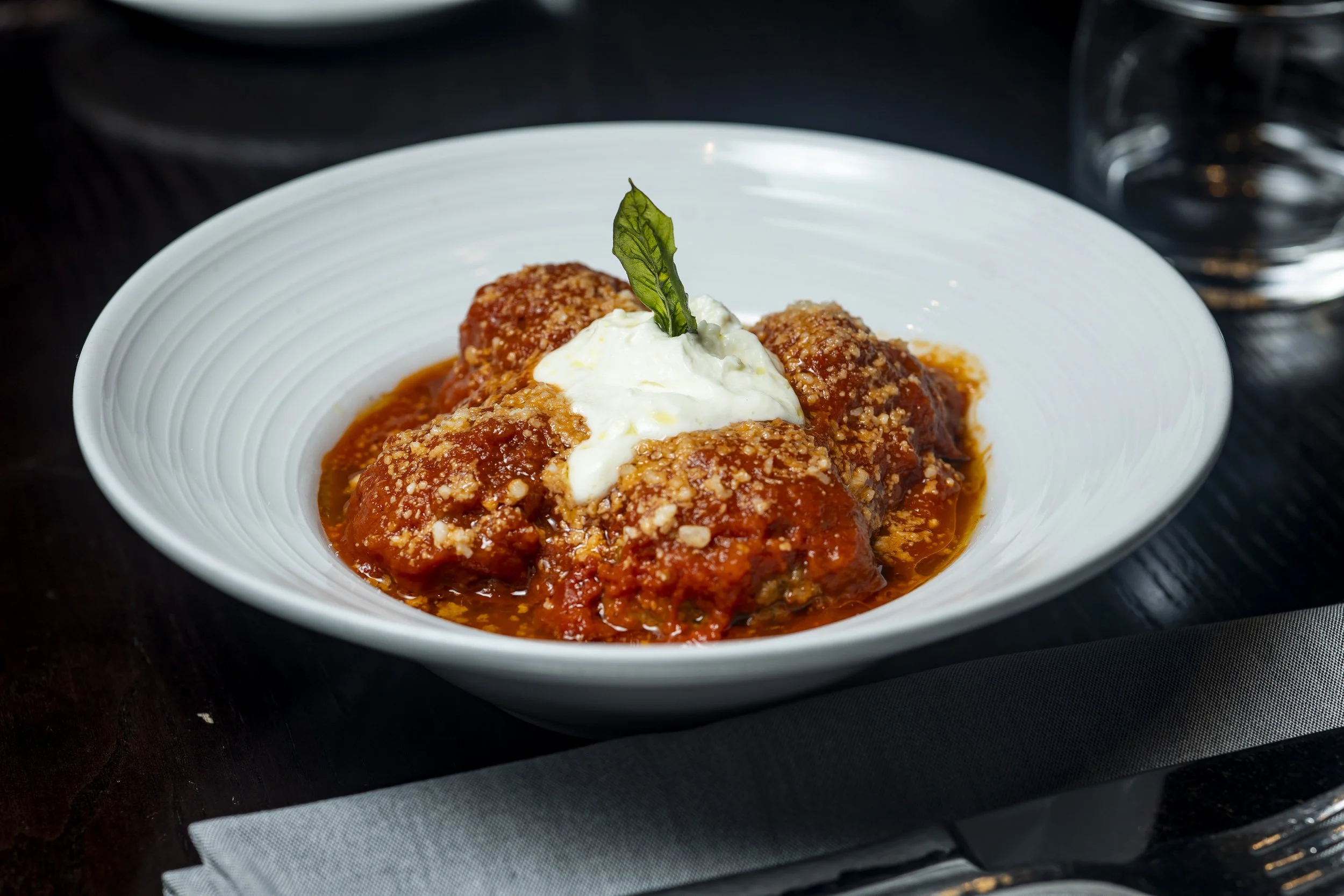 Meatballs with tomato sauce topped with cheese and a basil leaf in a white bowl on a dark table.