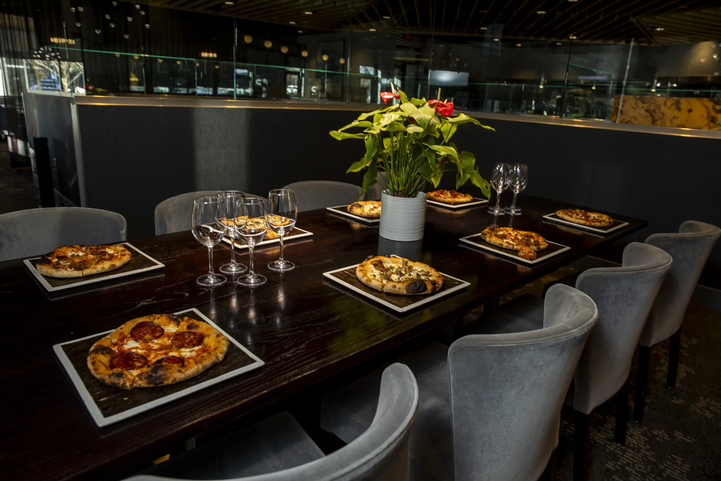 A dining table set for a pizza meal in a modern restaurant, with several plates of pizzas, empty wine glasses, and a potted plant as a centerpiece.
