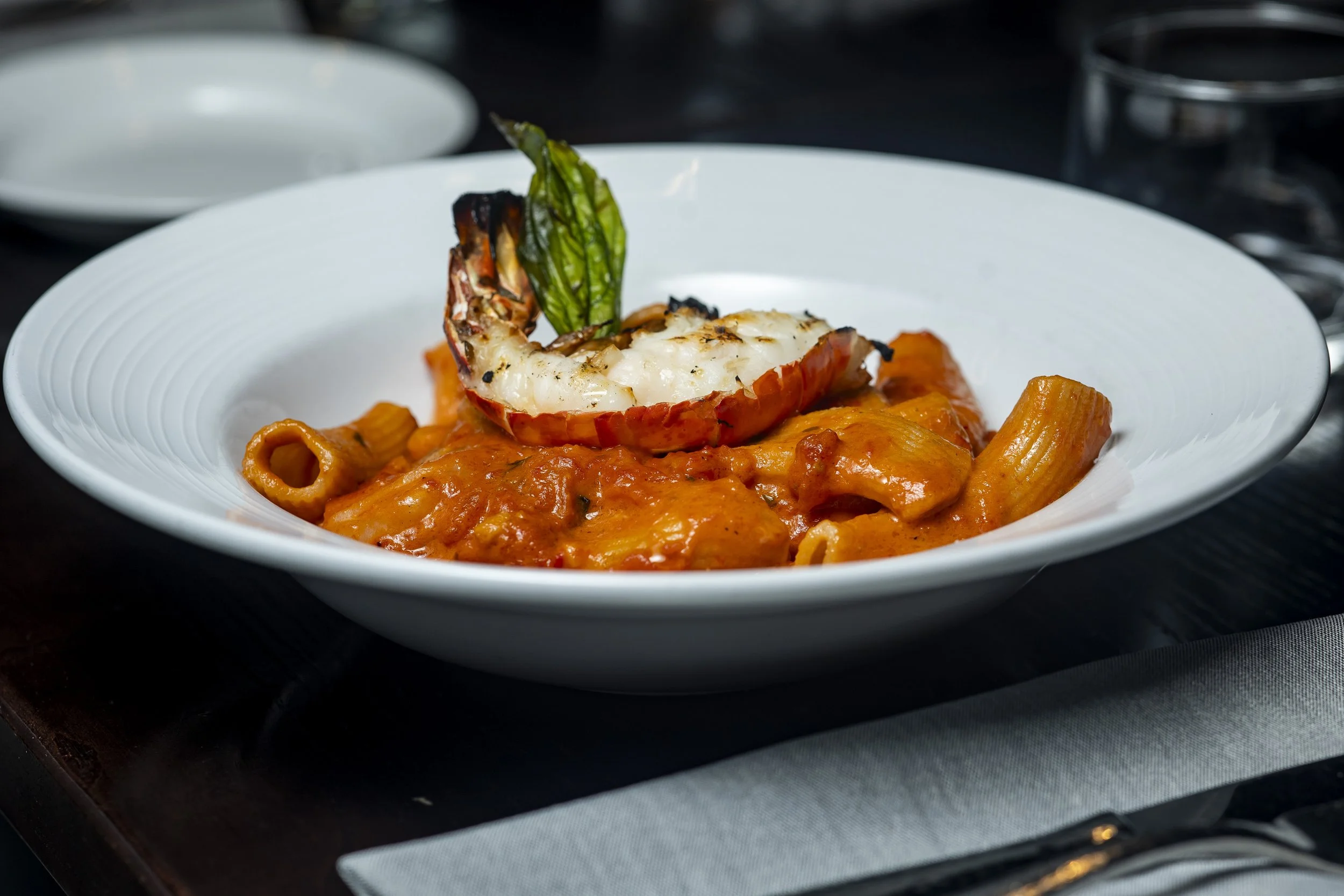 A white bowl of pasta with tomato sauce, topped with a grilled lobster tail and a grilled green vegetable garnished on top.