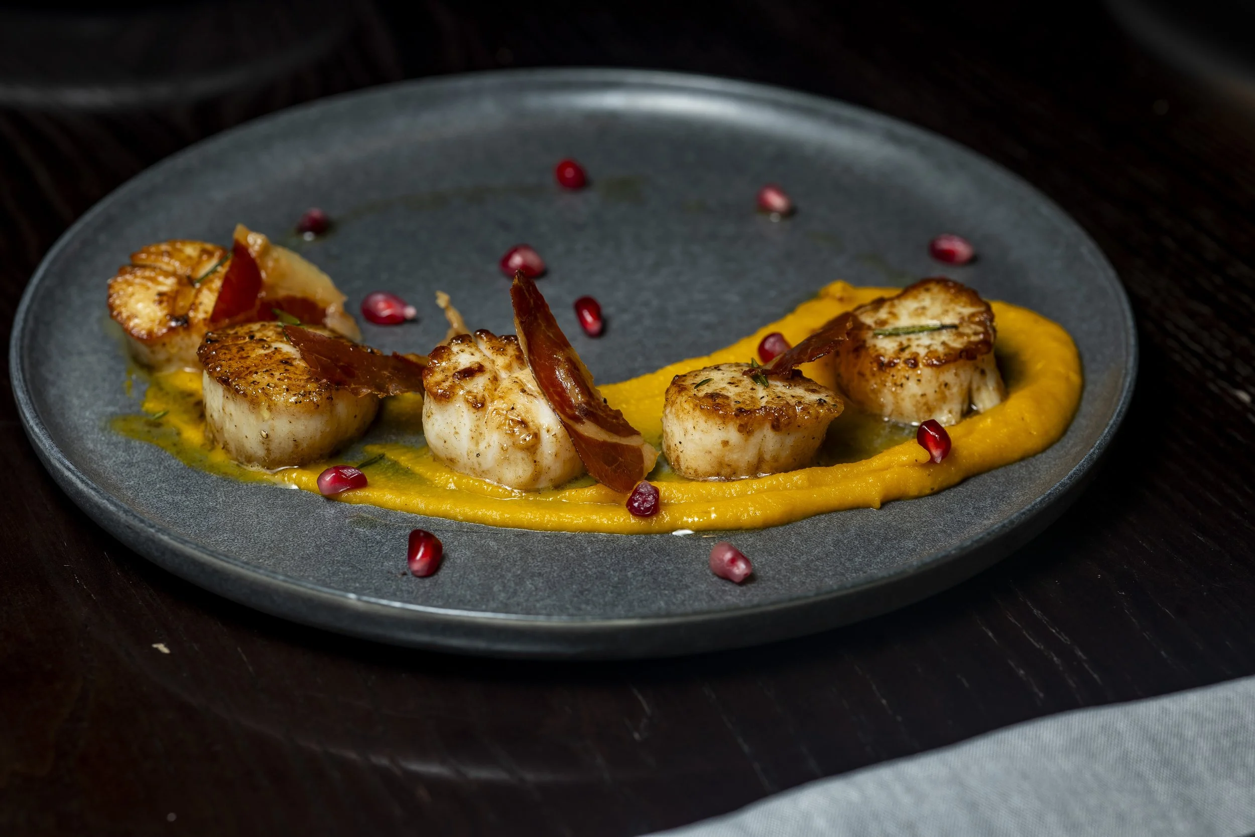 Seared scallops on a bed of orange puree, garnished with pomegranate seeds and thin slices of cured meat, served on a dark plate.