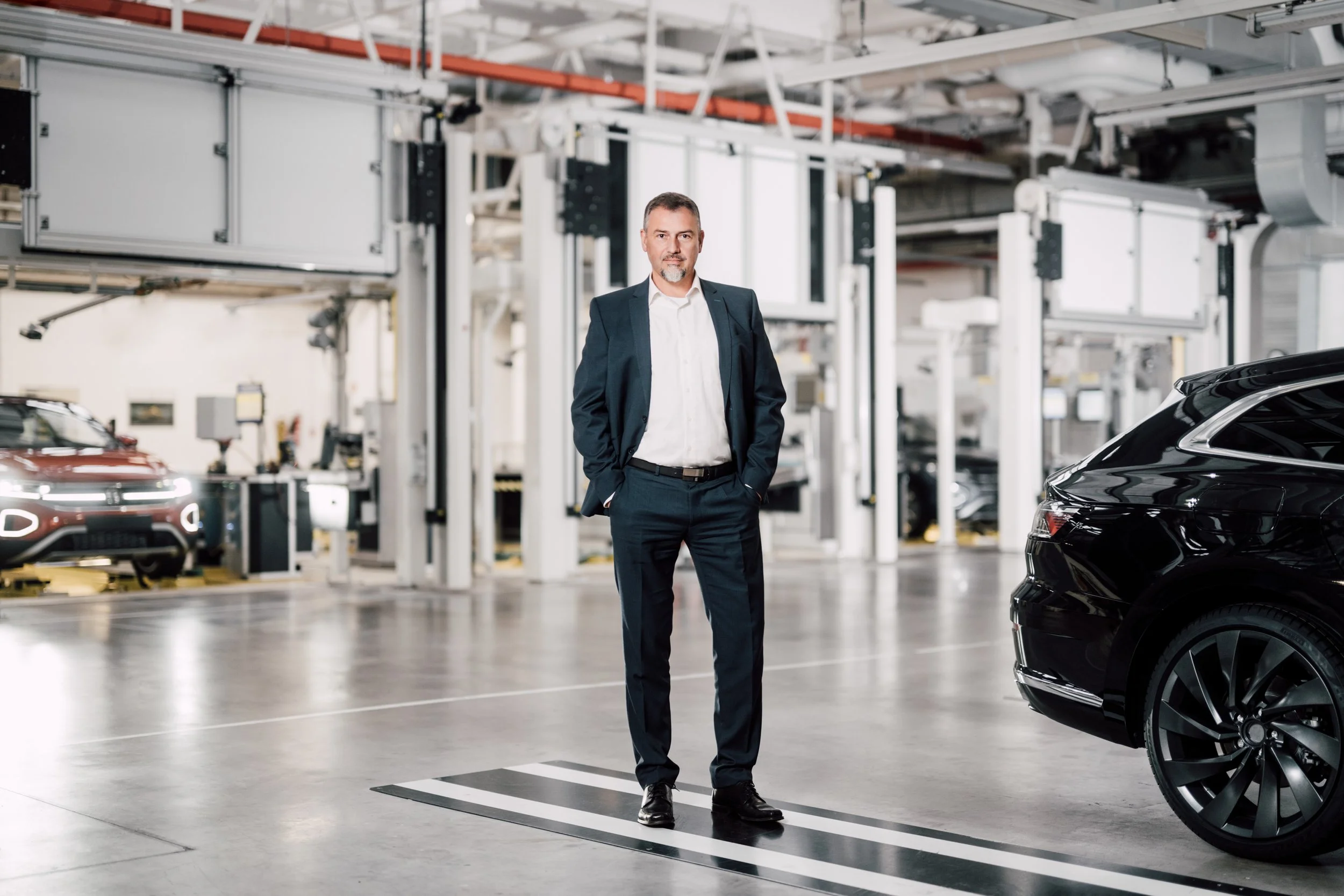 Ein Mann in Anzug steht in einer modernen Autofabrikhalle neben einem schwarzen Auto. Sehr qualitativ hochwertige Unternehmensfotografie.