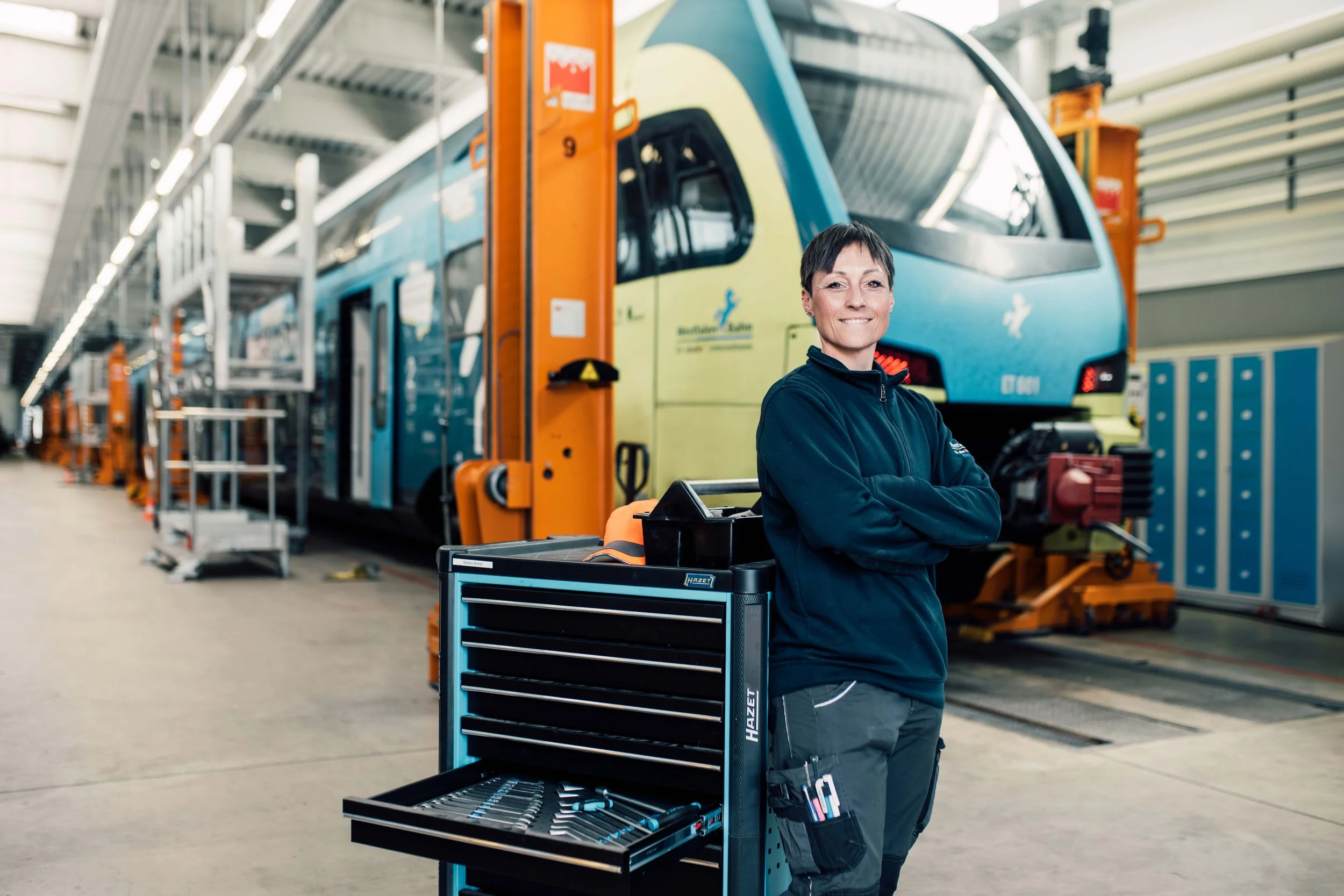 Eine Mitarbeiterin steht in einem Bahnbetriebshof mit ausgestatteter Werkzeugkiste vor einem modernen, blauen Zug. Es wirkt wie eine moderne Employer Branding Situation. Sie trägt eine schwarze Jacke und hat kurze dunkle Haare.