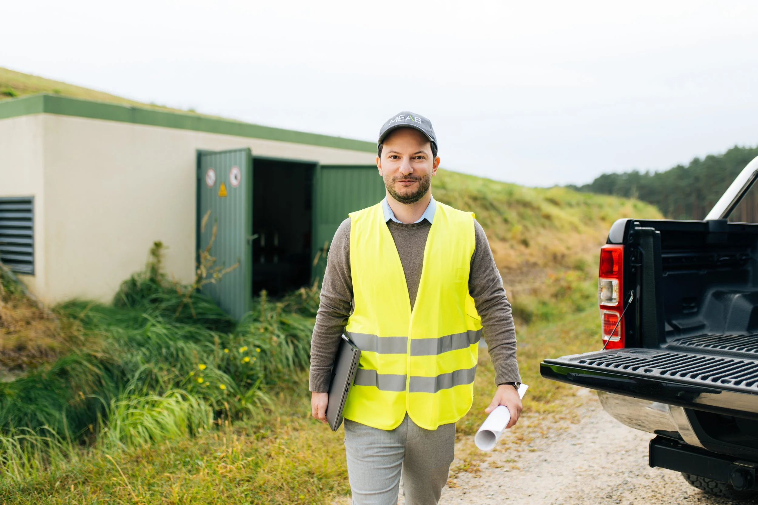 Mann in gelber Warnweste steht vor einem Gebäude im Freien, trägt einen Laptop und einen Rollladenausweis, hält eine Bauzeichnung in der Hand, neben einem weißen Pickup. Eine dynamische Employer Branding Kampagne.