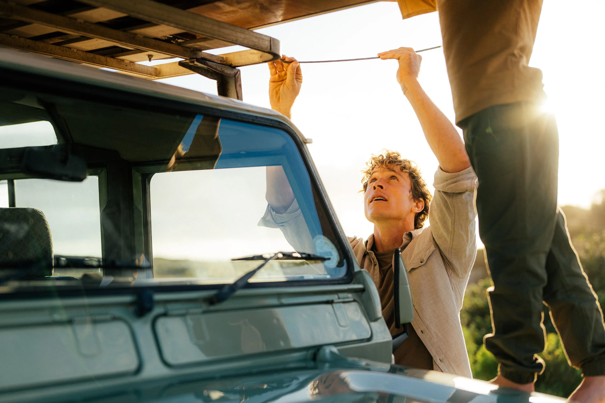 Zwei Menschen reparieren oder befestigen etwas an einem Dachträger auf einem alten Auto im Freien bei Sonnenuntergang.