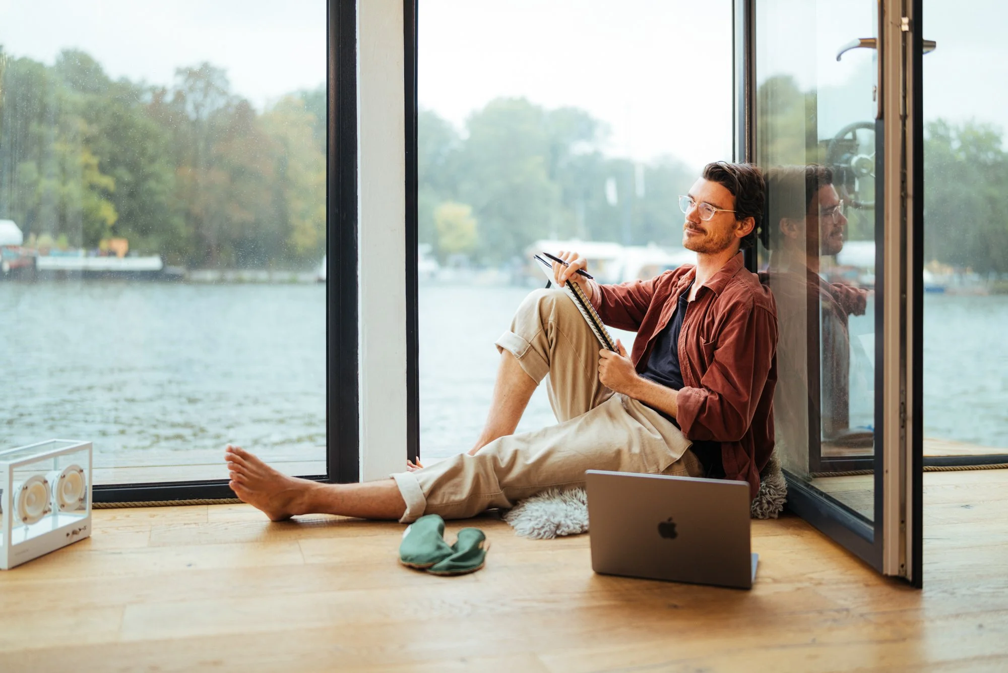 Ein Mann sitzt auf dem Boden vor einer großen Fensterscheibe mit Blick auf Wasser und Bäume, schreibt in ein Notizbuch, hat einen Laptop vor sich und trägt Brille und eine braune Jacke. Eine besondere Homeoffice Bildserie!