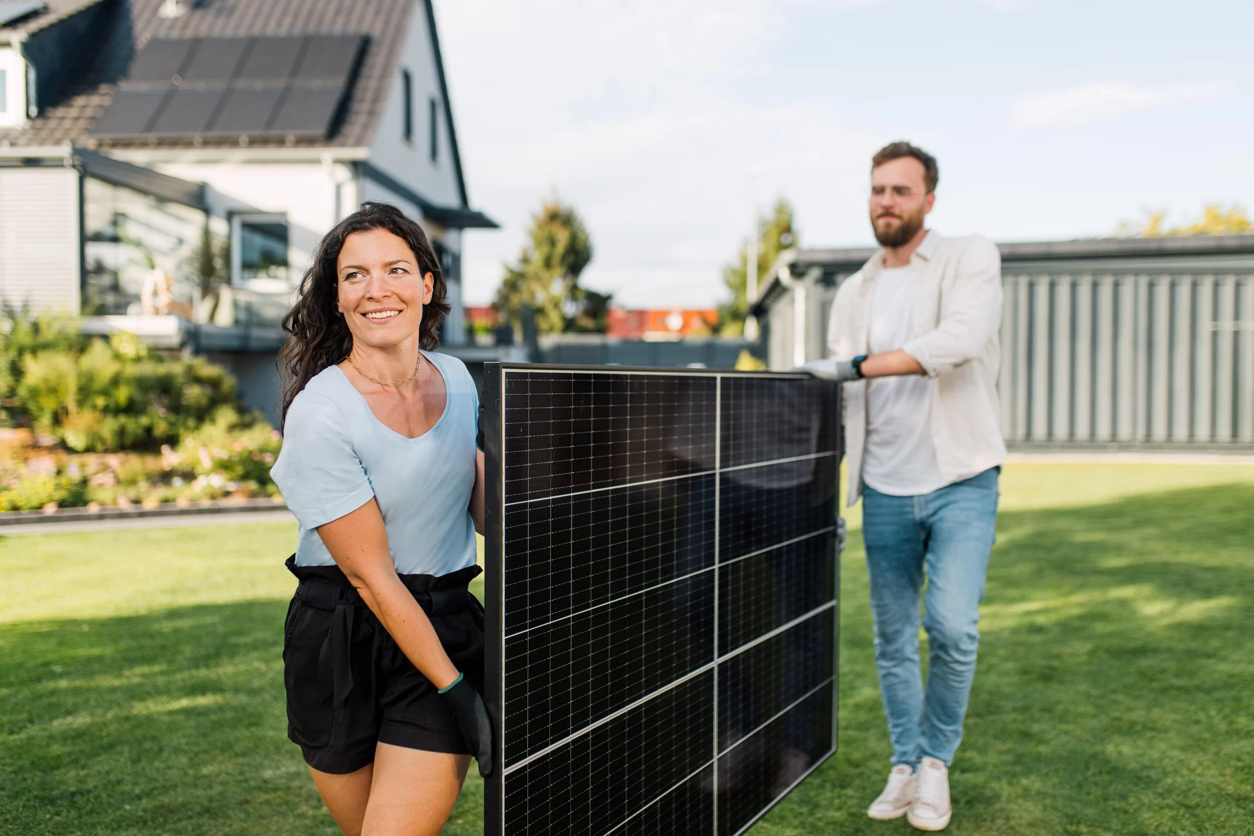 Zwei Personen,ein Mann und eine Frau, installiere Solarzellen auf einer Wiese im Garten. Eine Werbekampagne für Solarmodule. Der Himmel ist blau und das Haus im Hintergrund hat Solarpaneele auf dem Dach.