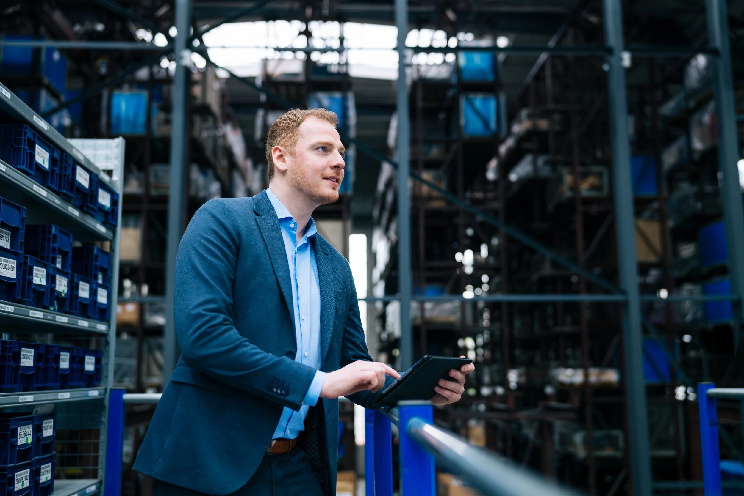 Junger Mann in Anzug mit Tablet in einem Lagerhaus mit Metallregalen voll mit Kartons. Corporate Fotografie aus dem Bilderbuch.