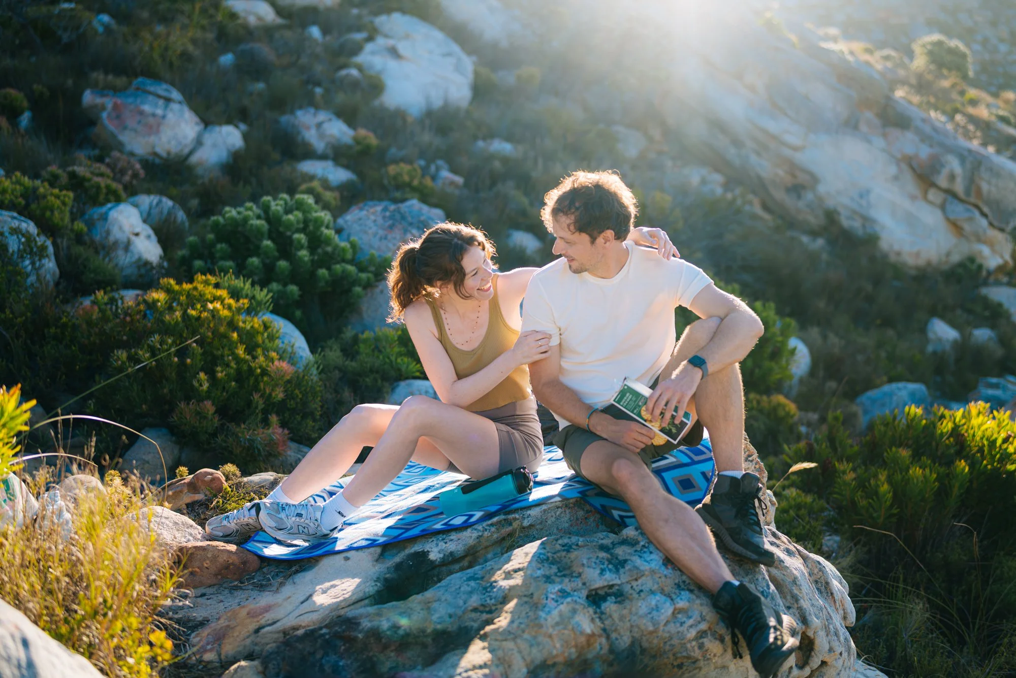 Ein Mann und eine Frau sitzen auf einem Felsen in einer bergigen Landschaft bei Sonnenuntergang, lachen und genießen die gemeinsame Zeit.