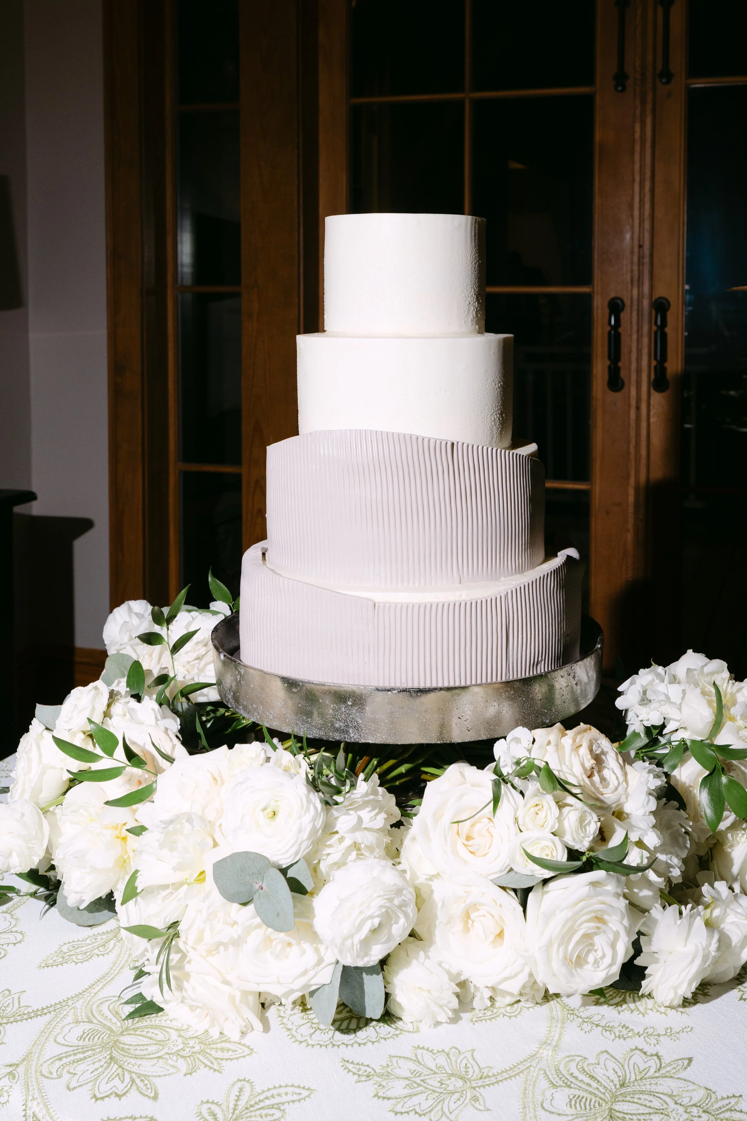 A four-tier white wedding cake with textured icing, surrounded by white flowers and greenery, placed on a table with a decorative cloth in a dimly lit room.