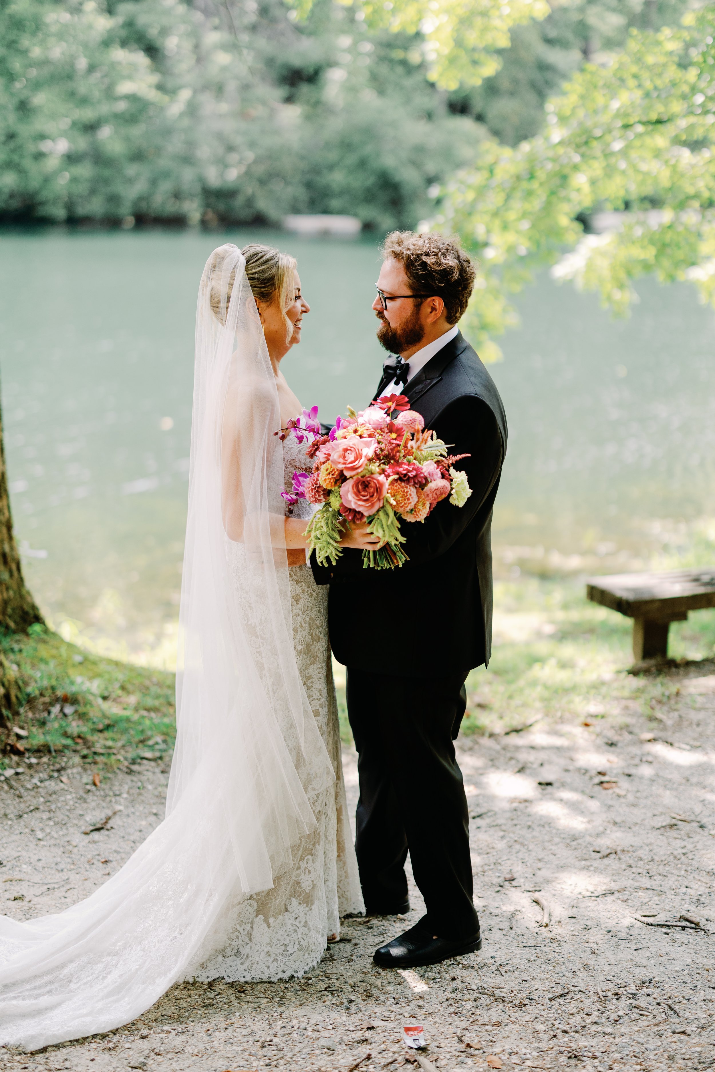A bride and groom standing outdoors near a body of water, holding a colorful bouquet, gazing at each other on their wedding day.