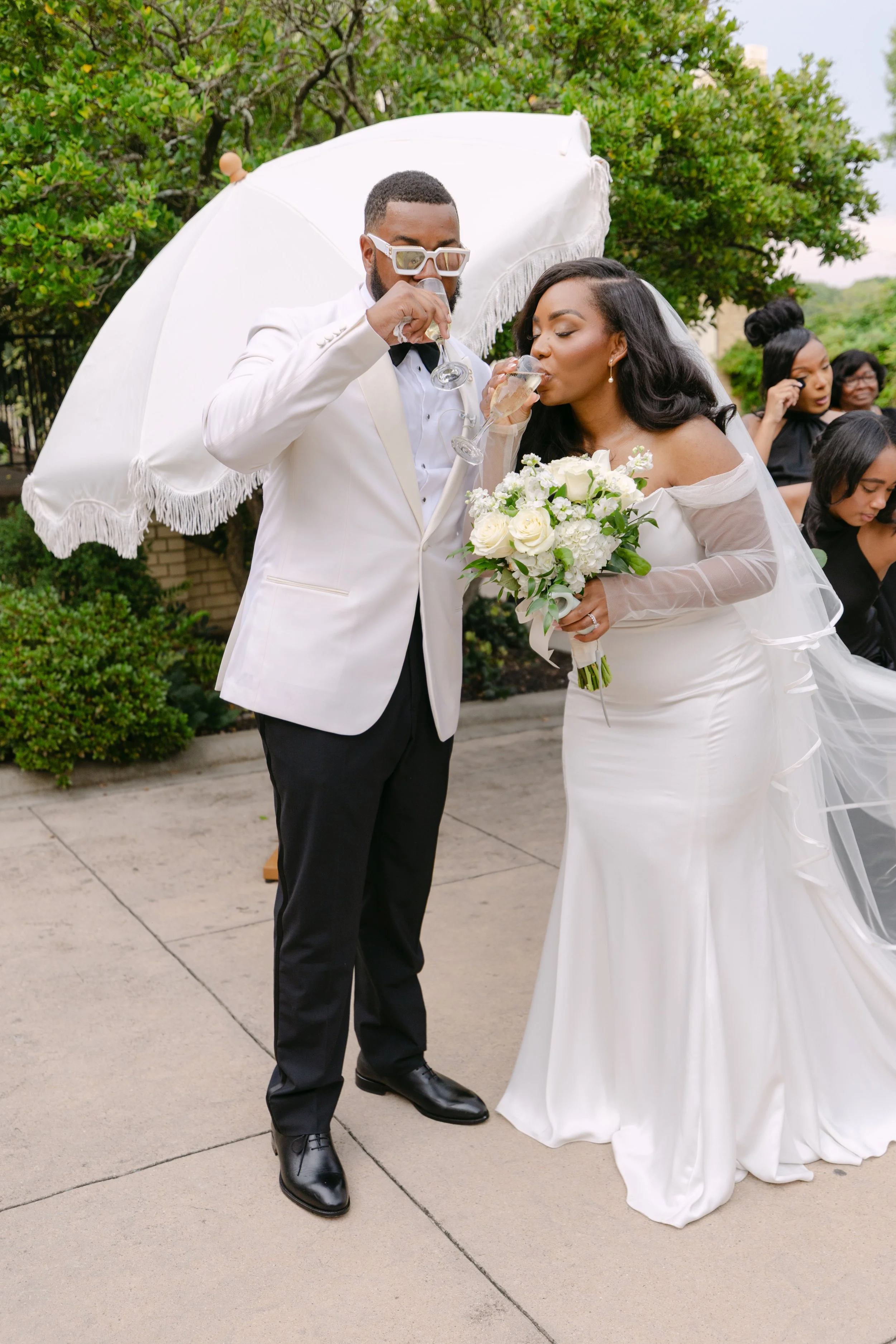 A newlywed couple dressed in wedding attire, the groom in a white tuxedo and the bride in a white wedding gown, are drinking champagne from glasses outdoors under a white parasol. The bride holds a bouquet of white roses. In the background, a group o