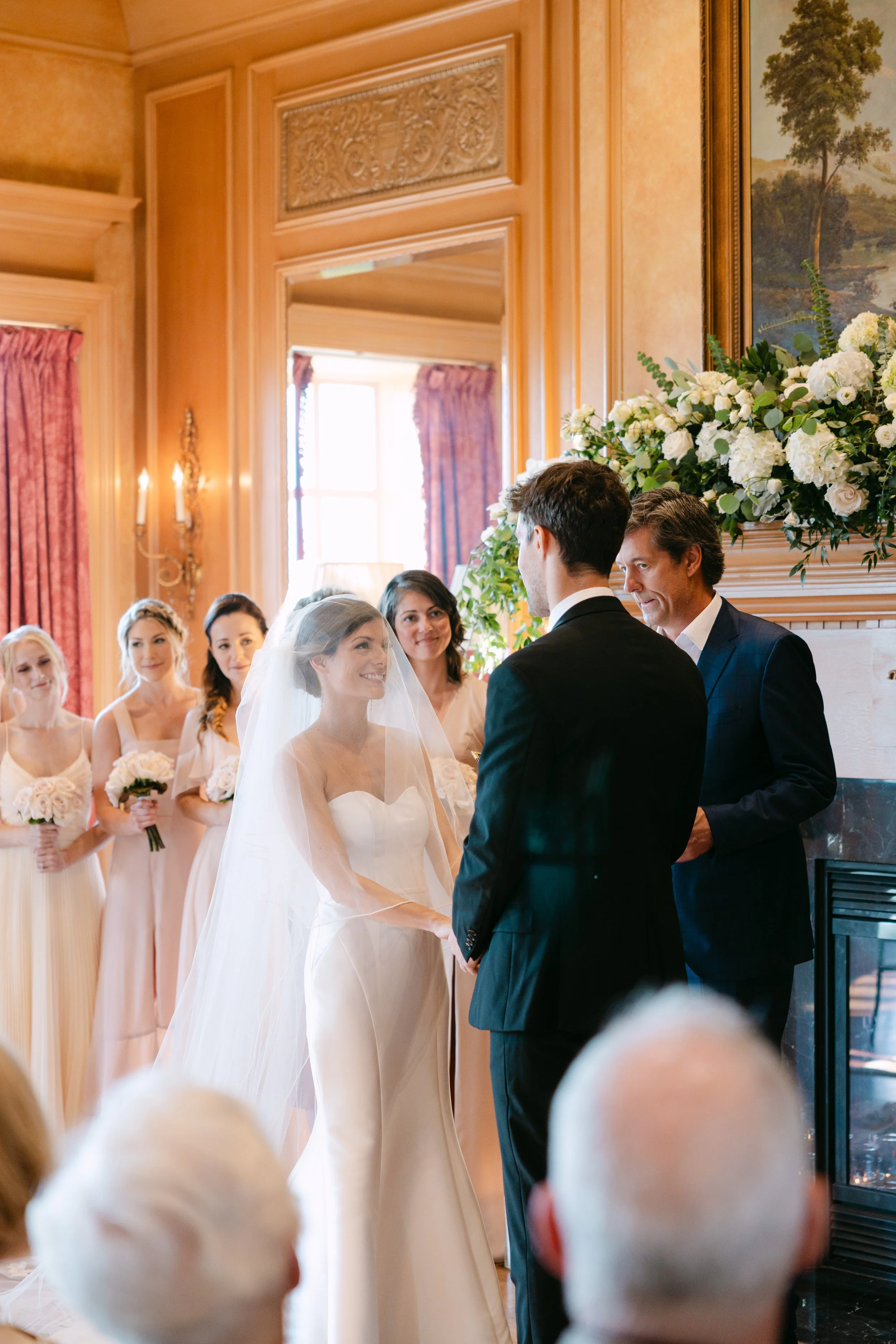 A bride and groom exchanging vows during a wedding ceremony in an elegant, wood-paneled room with a fireplace decorated with a large floral arrangement, and a bridal party of bridesmaids in pastel dresses standing behind them.