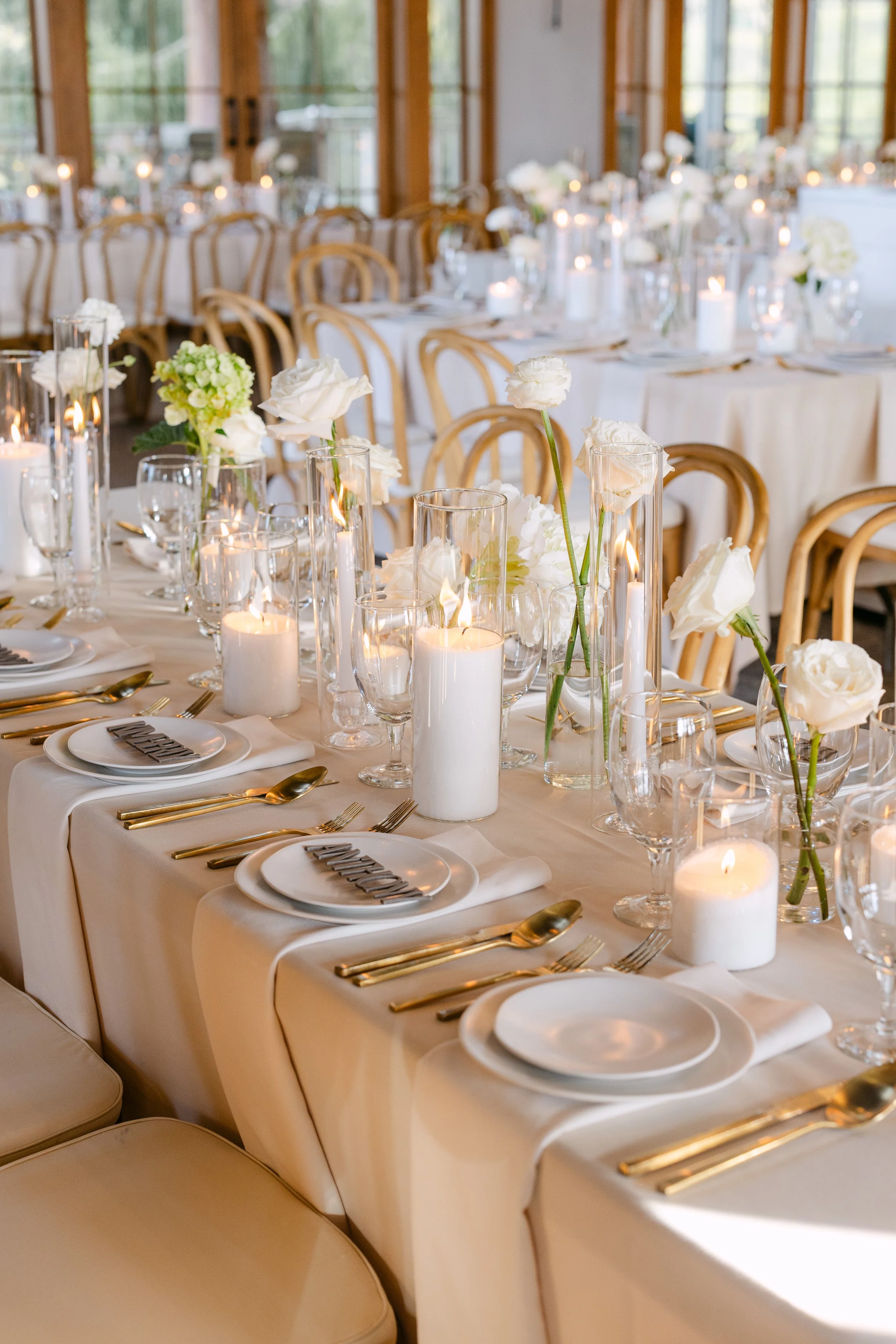 Elegant wedding reception table setup with white flowers, white candles, and gold cutlery on a cream-colored tablecloth.