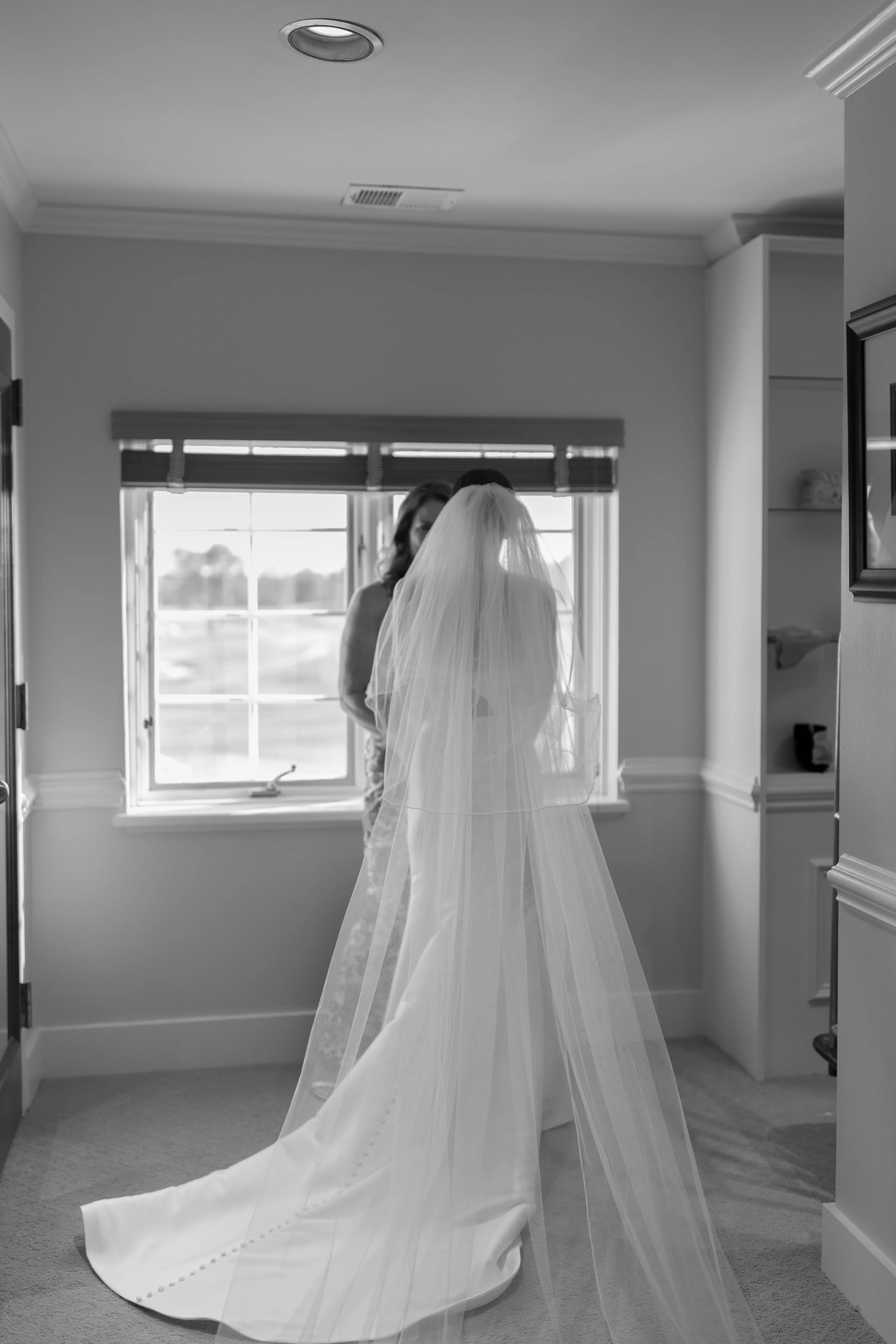 A bride standing in a room with large window, wearing a wedding dress and veil, facing another woman who appears to be helping her get ready.
