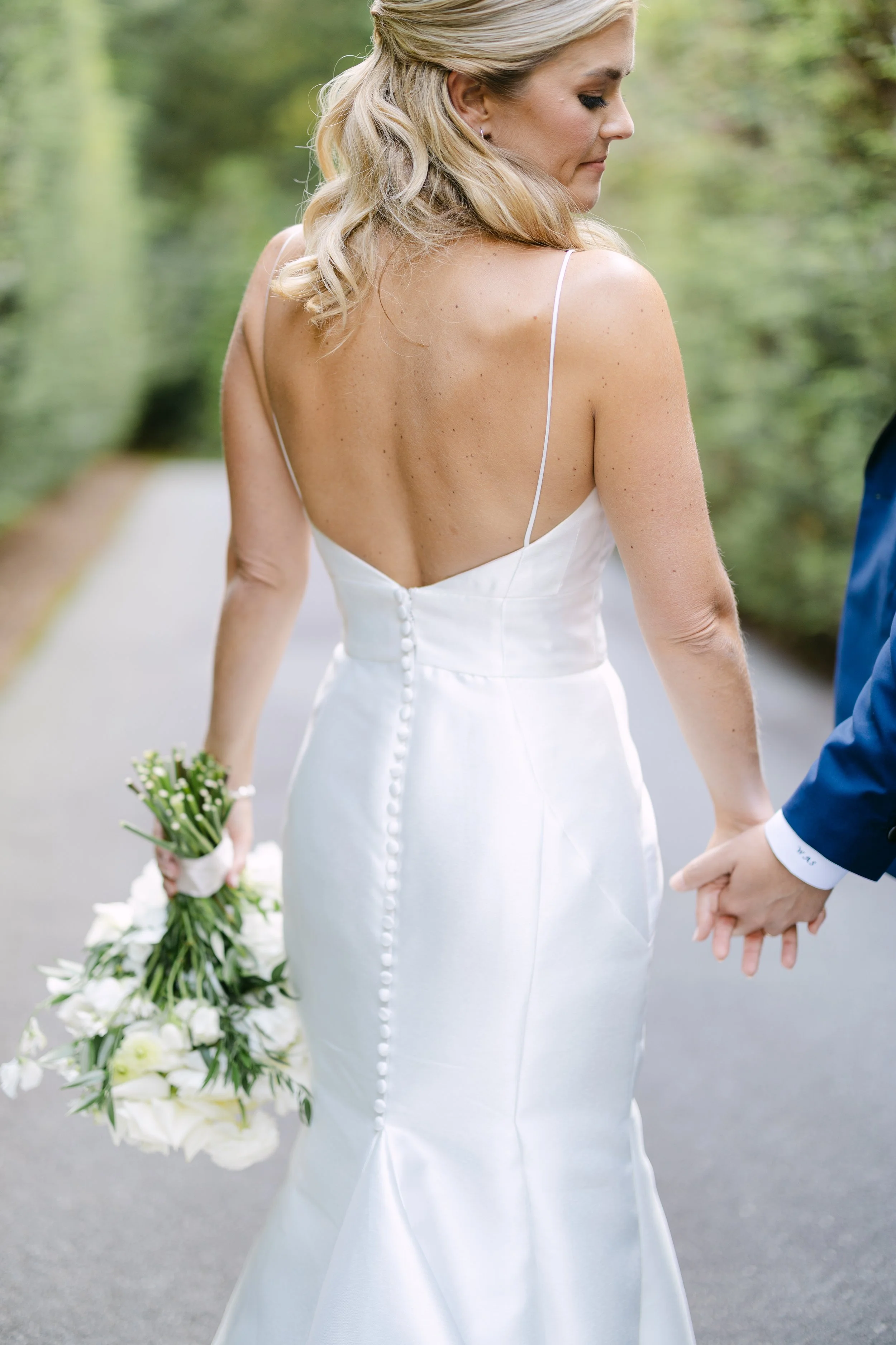 Bride in a white wedding dress holding a bouquet of white flowers, standing next to a groom on a path surrounded by greenery.