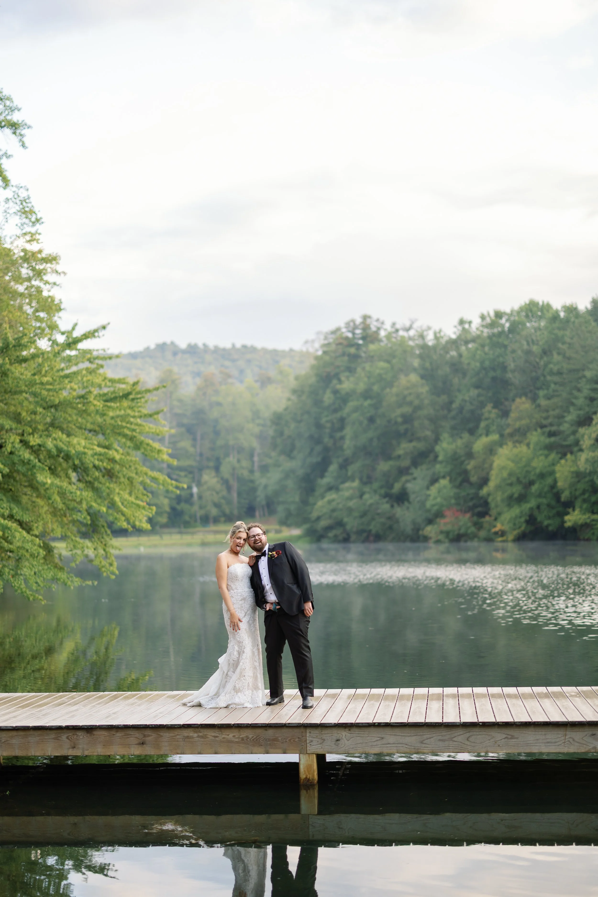 Bride and groom standing on a wooden dock by a lake, surrounded by green trees and distant hills, smiling and posing for their wedding photo.