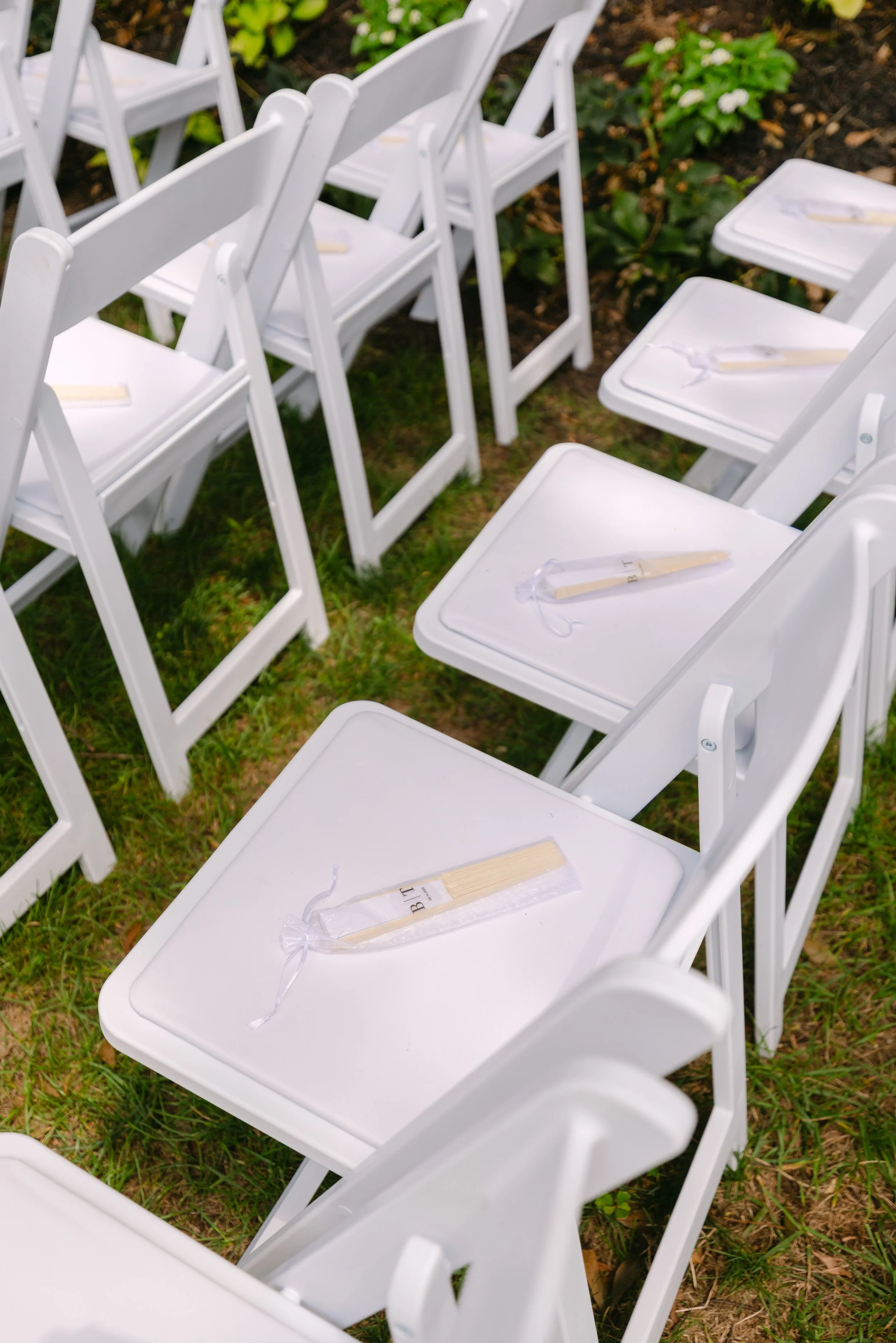 White folding chairs arranged outdoors on grass, each with a white ribbon-tied favor on the seat.