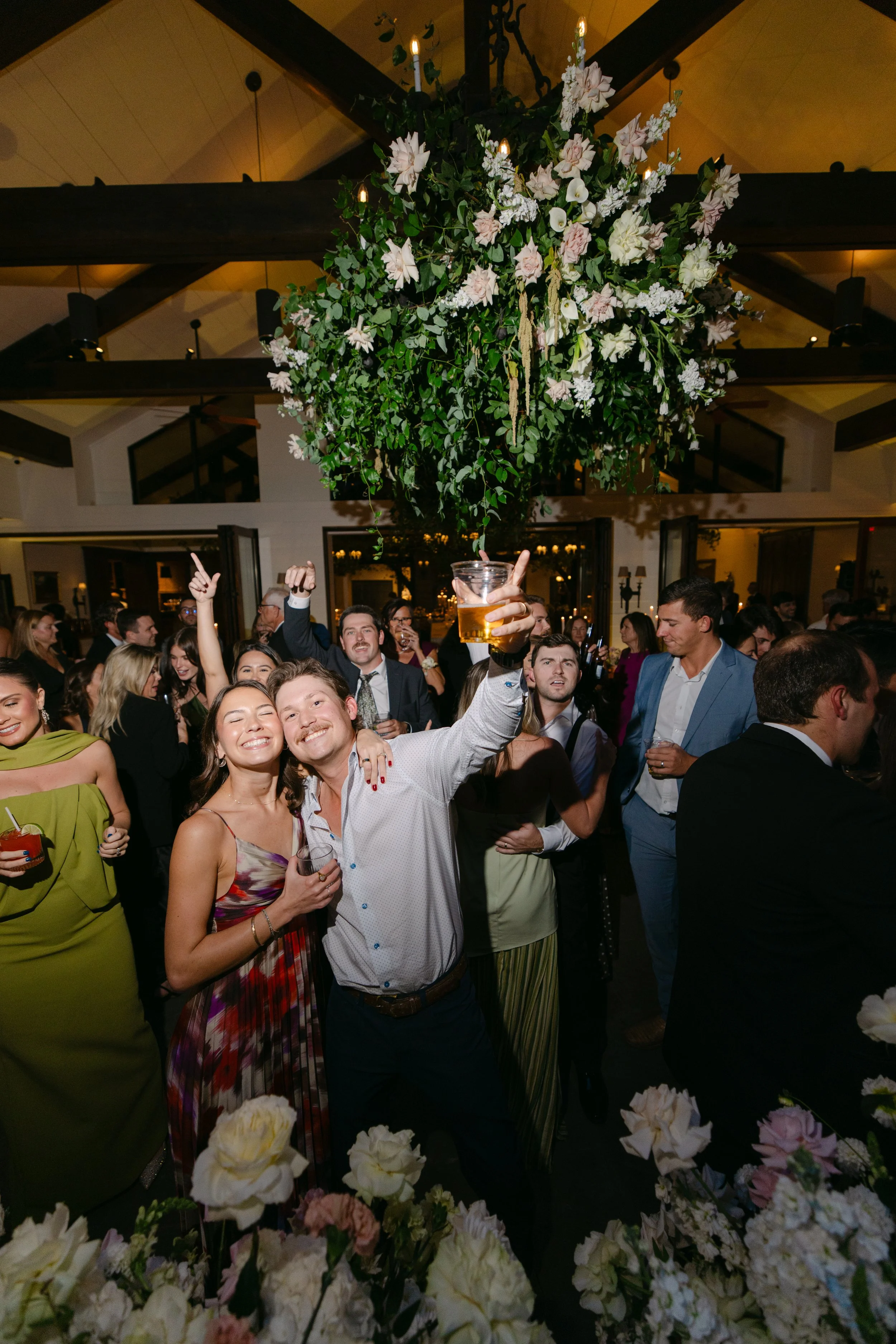 Crowded wedding reception with guests raising drinks beneath a large floral arrangement and candlelit timber-beam ceiling