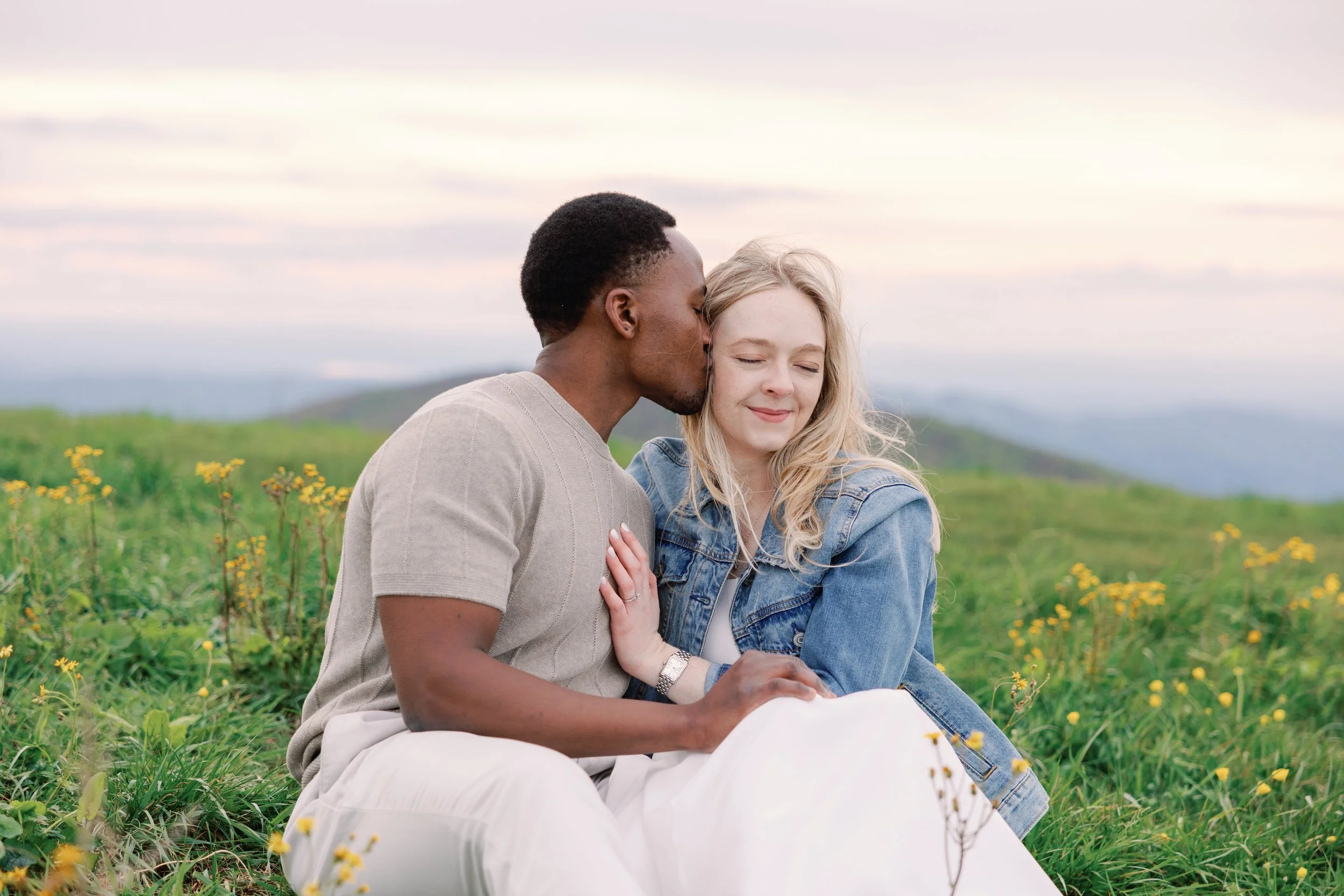 A couple sitting in a field of yellow flowers with a scenic landscape and cloudy sky in the background. The man is giving the woman a kiss on the forehead, and she has a gentle smile with her eyes closed.