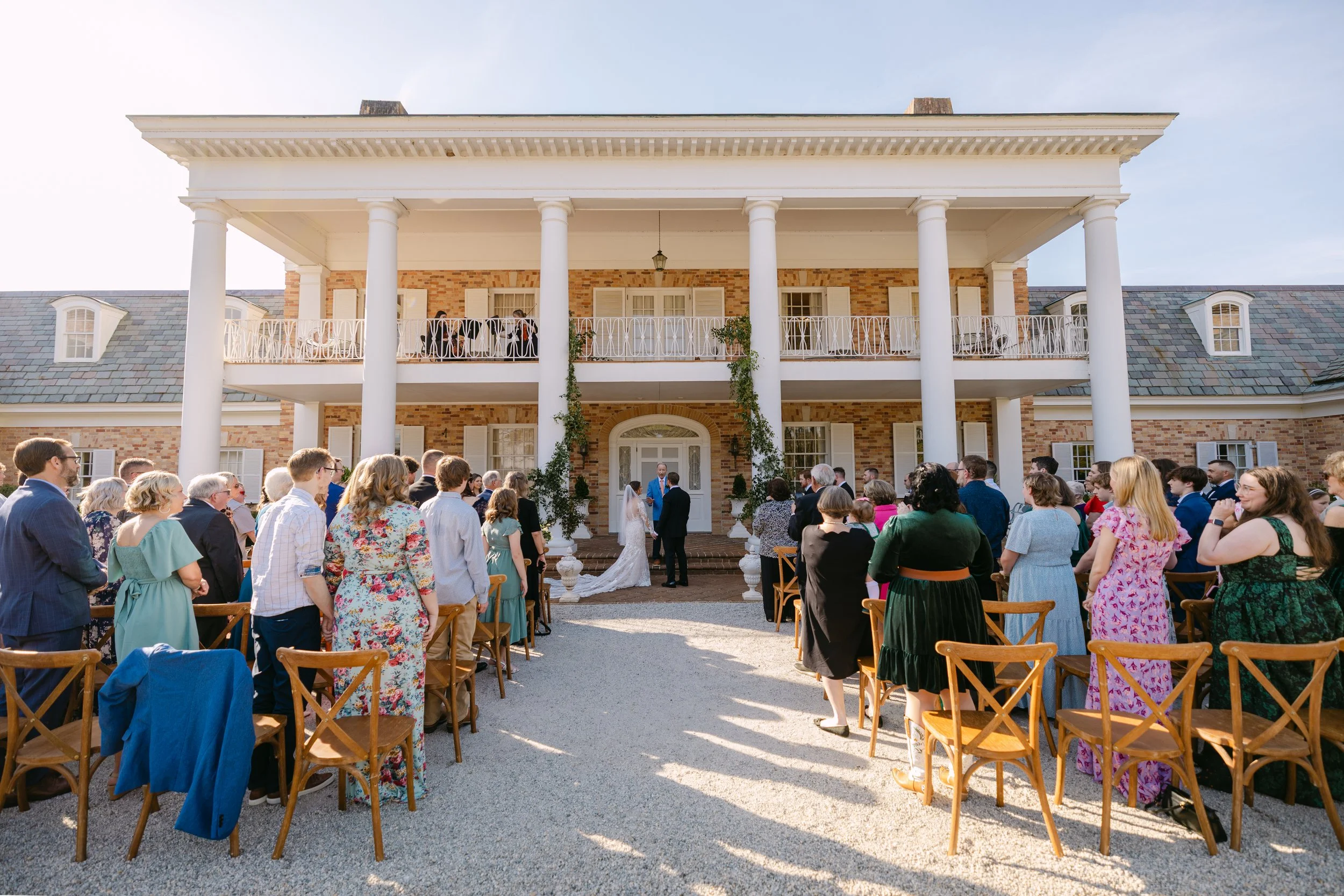 Wedding ceremony taking place outside a large brick house with white pillars, where the bride and groom stand before officiant amidst seated guests.