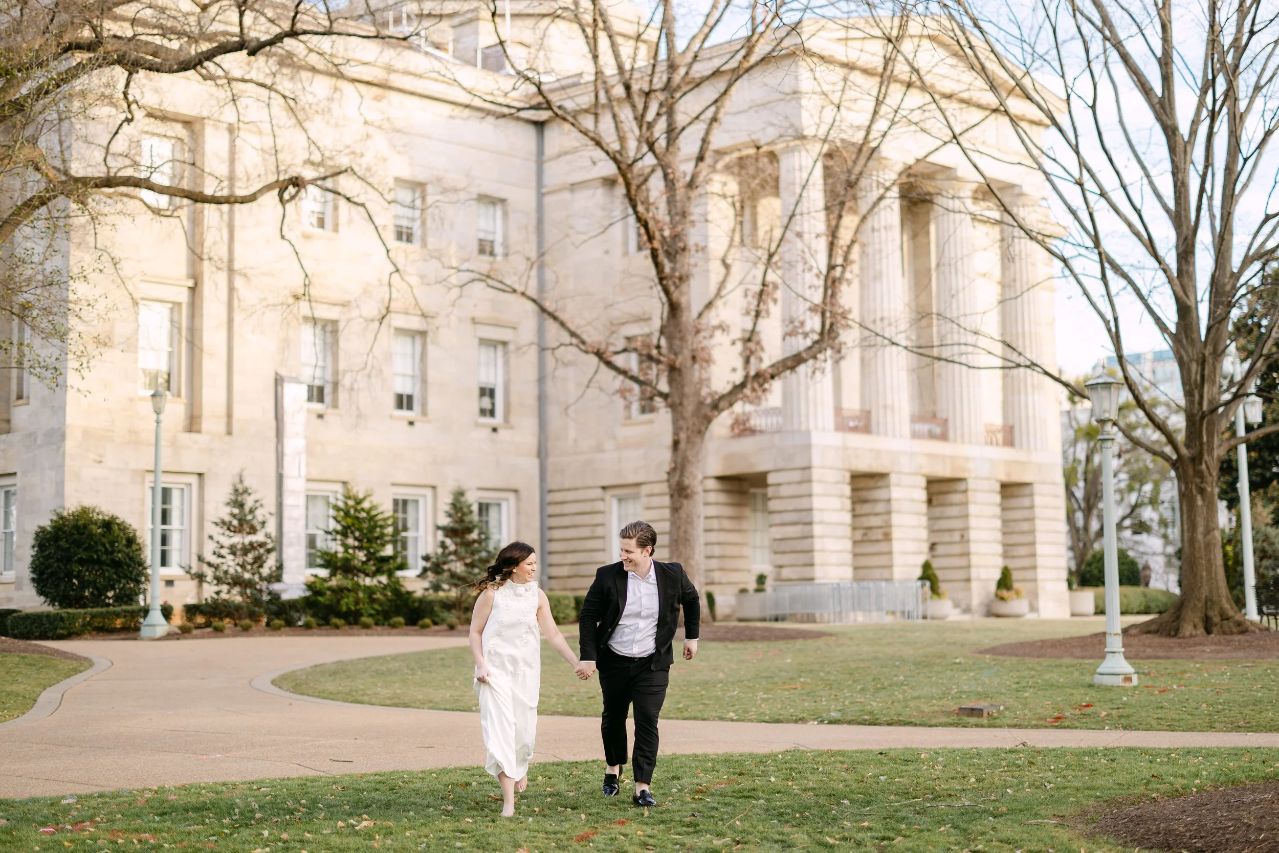 A couple walking hand in hand on a park pathway in front of a large historic building with columns, trees, grass, and lampposts.