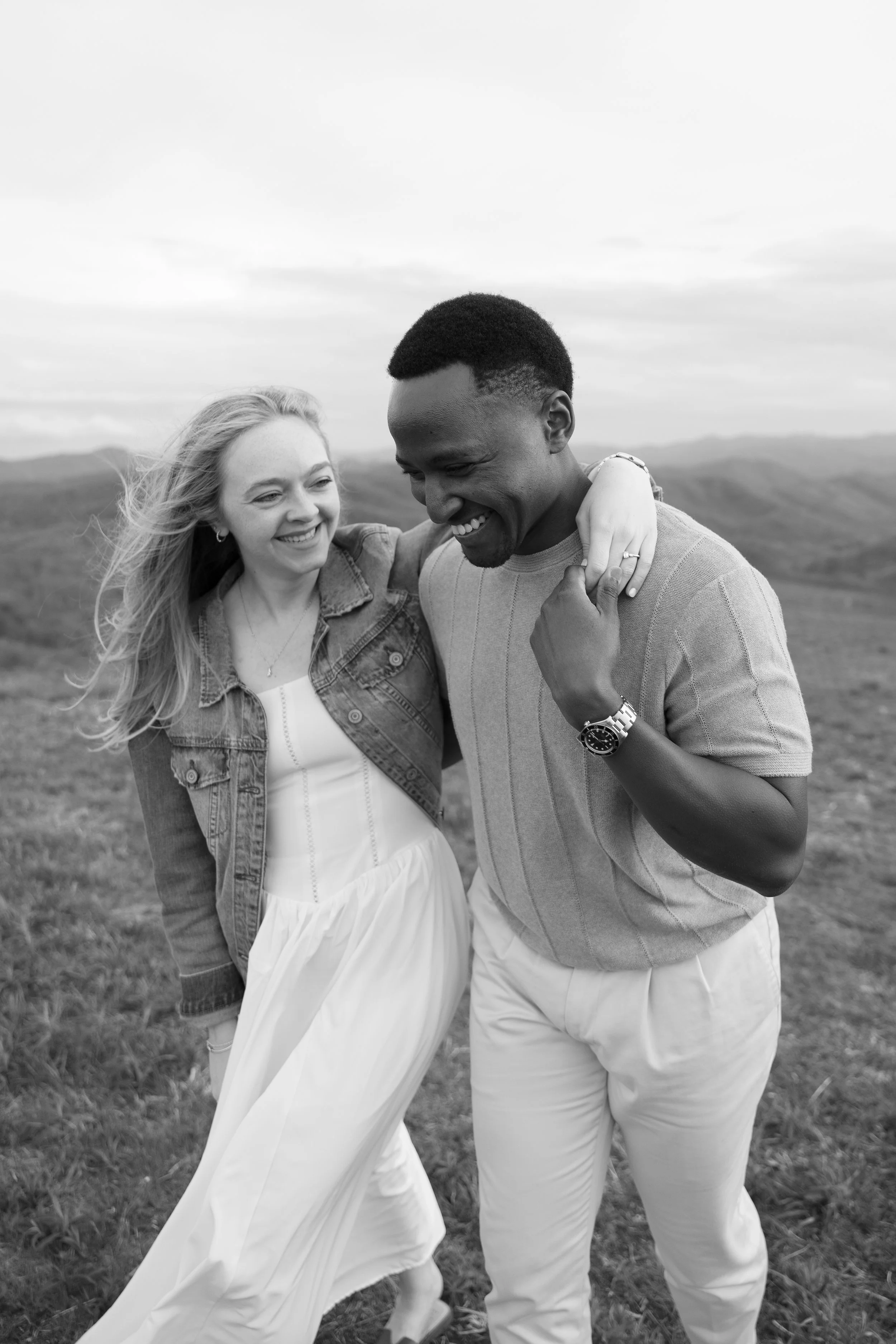 A smiling woman and man standing outdoors on a grassy field, embracing each other.
