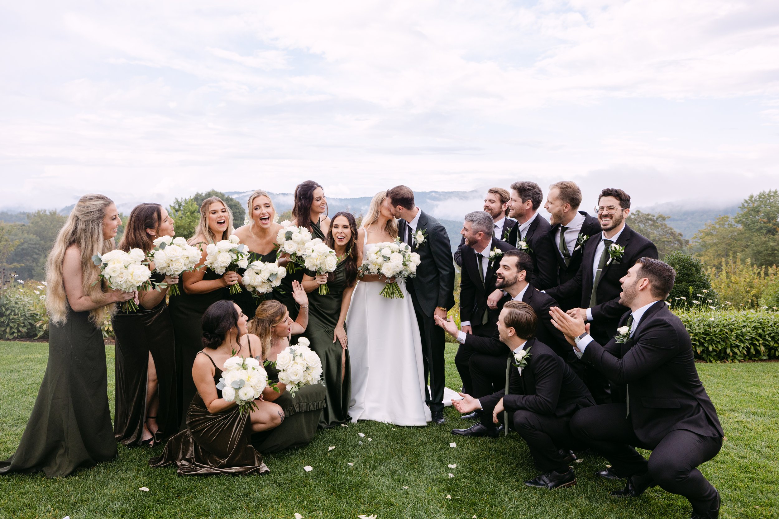 A wedding celebration outdoors with a bride and groom kissing, surrounded by bridesmaids and groomsmen holding bouquets and wearing formal attire