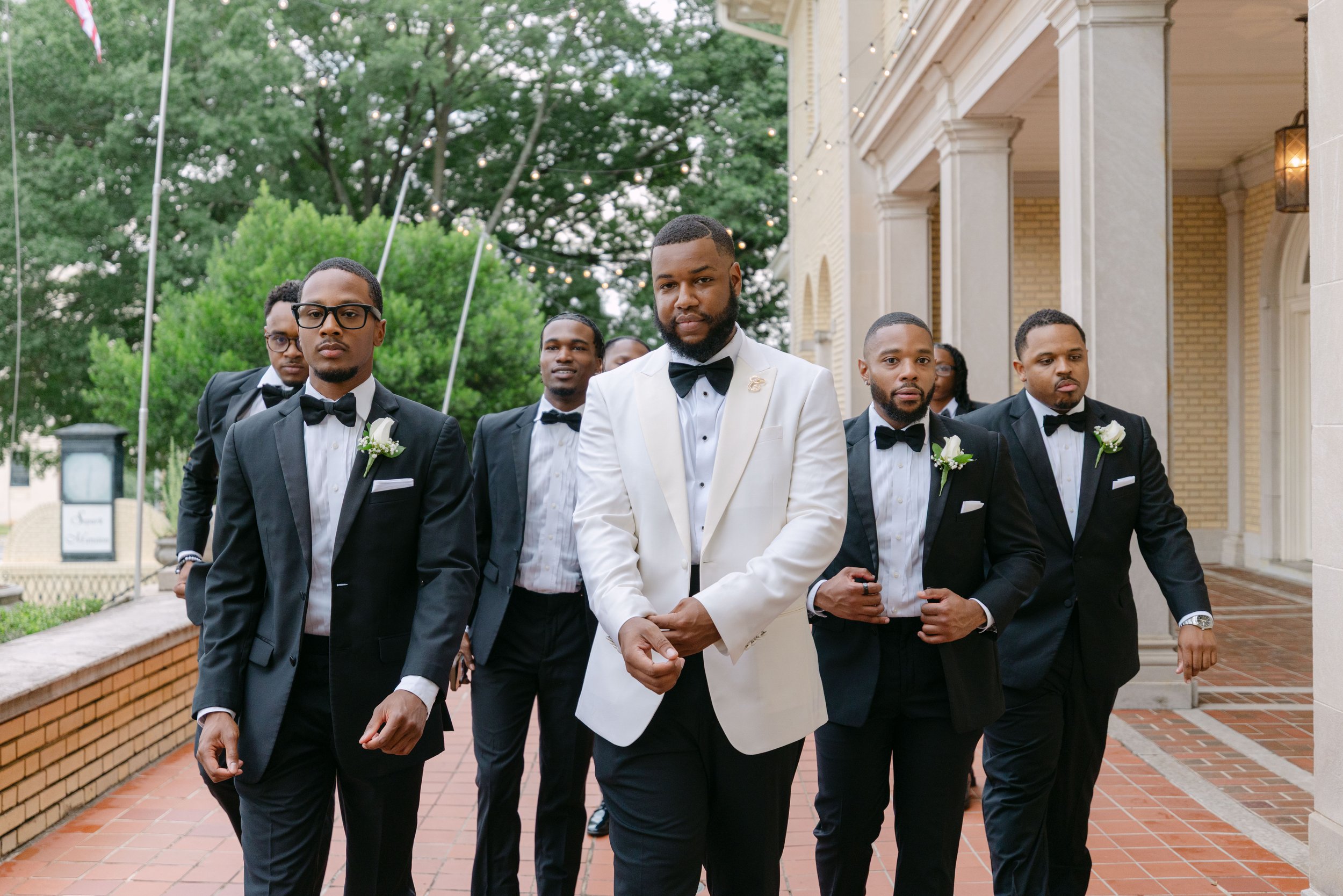 A group of six men dressed in formal tuxedos, some with bow ties and boutonnières, walking outdoors near a building with string lights and trees in the background.