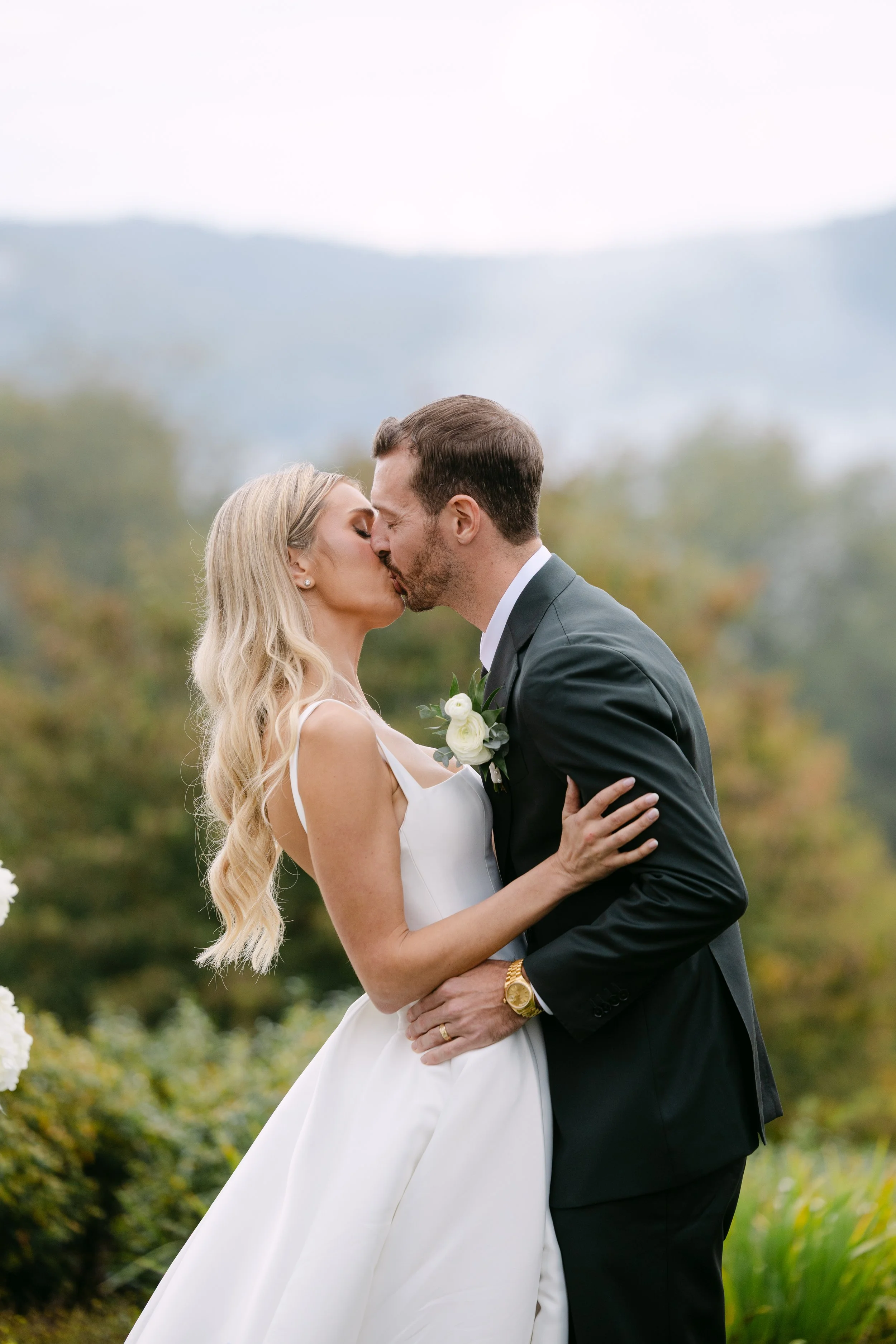 A bride and groom share a kiss outdoors during their wedding ceremony, with a blurred natural landscape in the background.