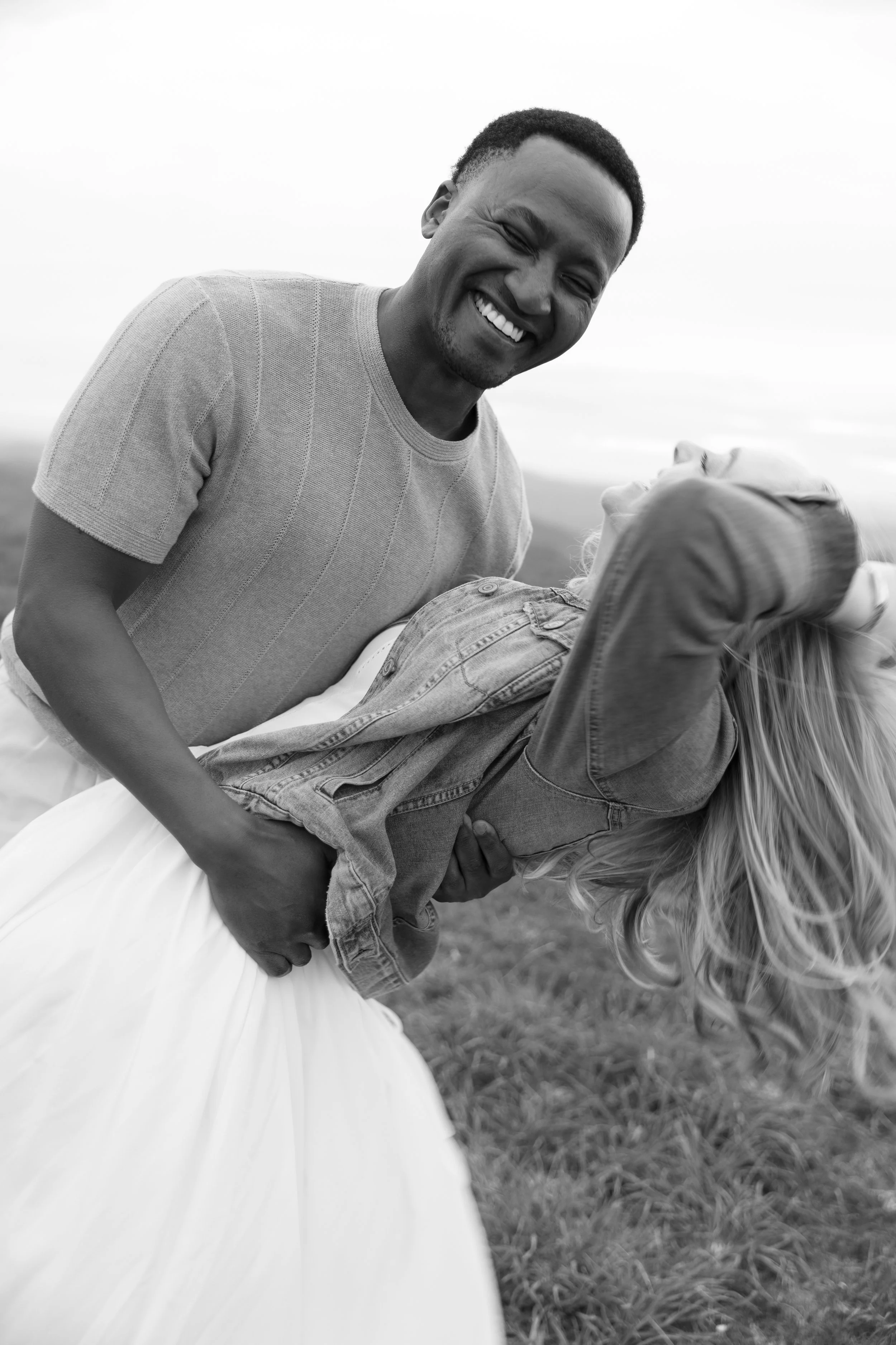 A man with a short haircut smiling, holding a woman in a dance pose outdoors on grass, with a blurred sky background, in black and white.