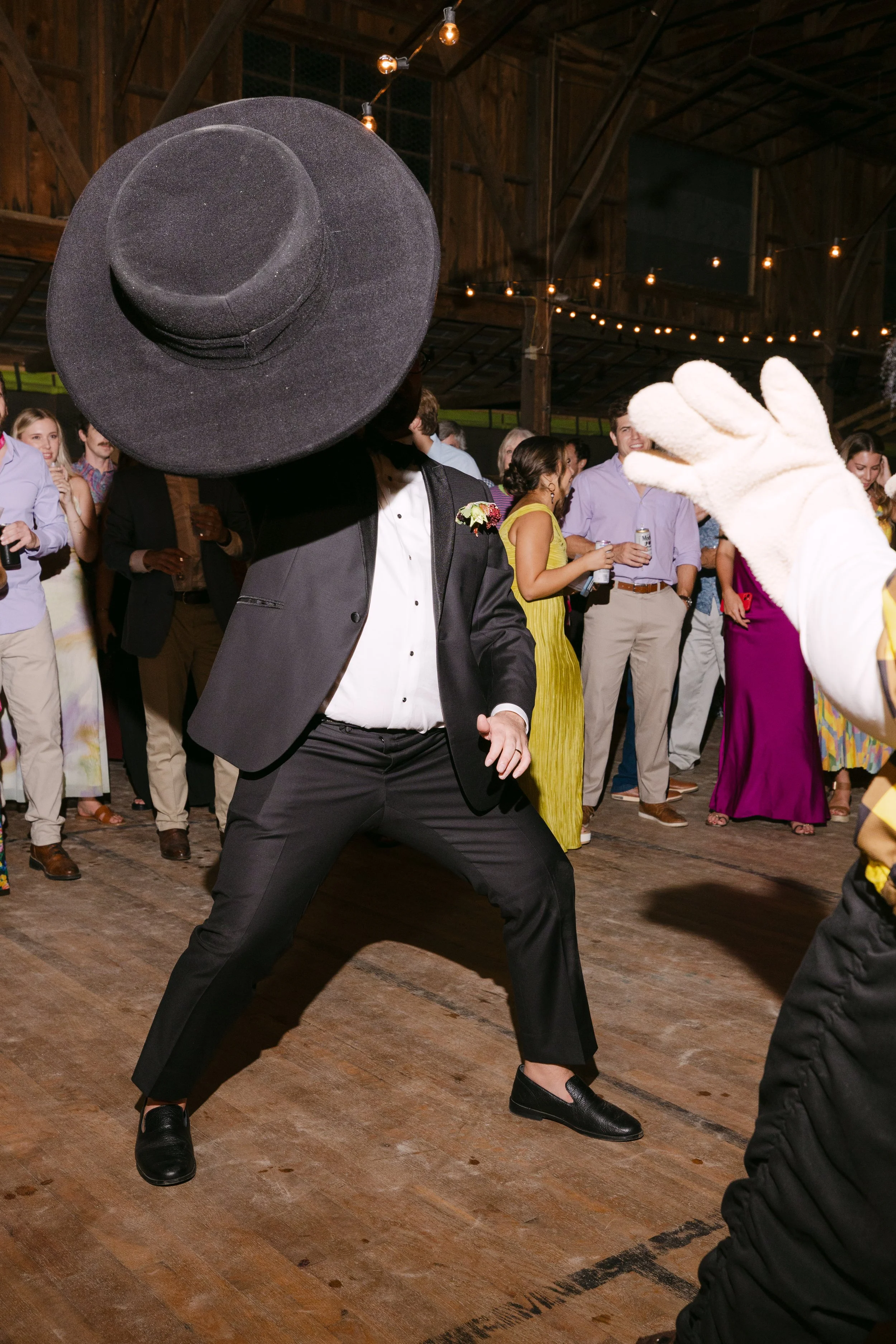 Person in black suit and large black hat dancing at a party with a group of people in the background.