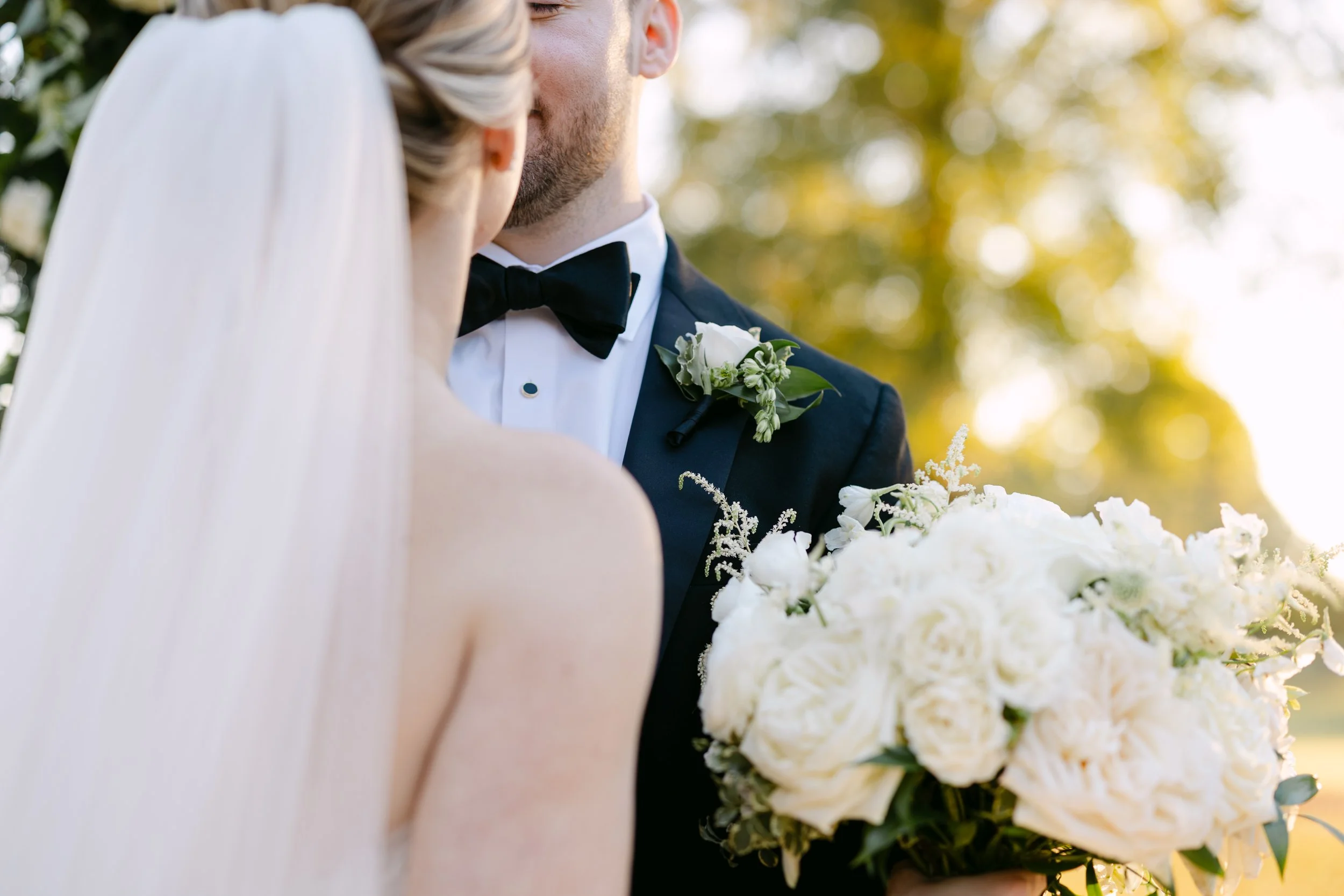 Bride and groom kissing outdoors, groom holding a bouquet of white roses, with sunlight and trees in the background.